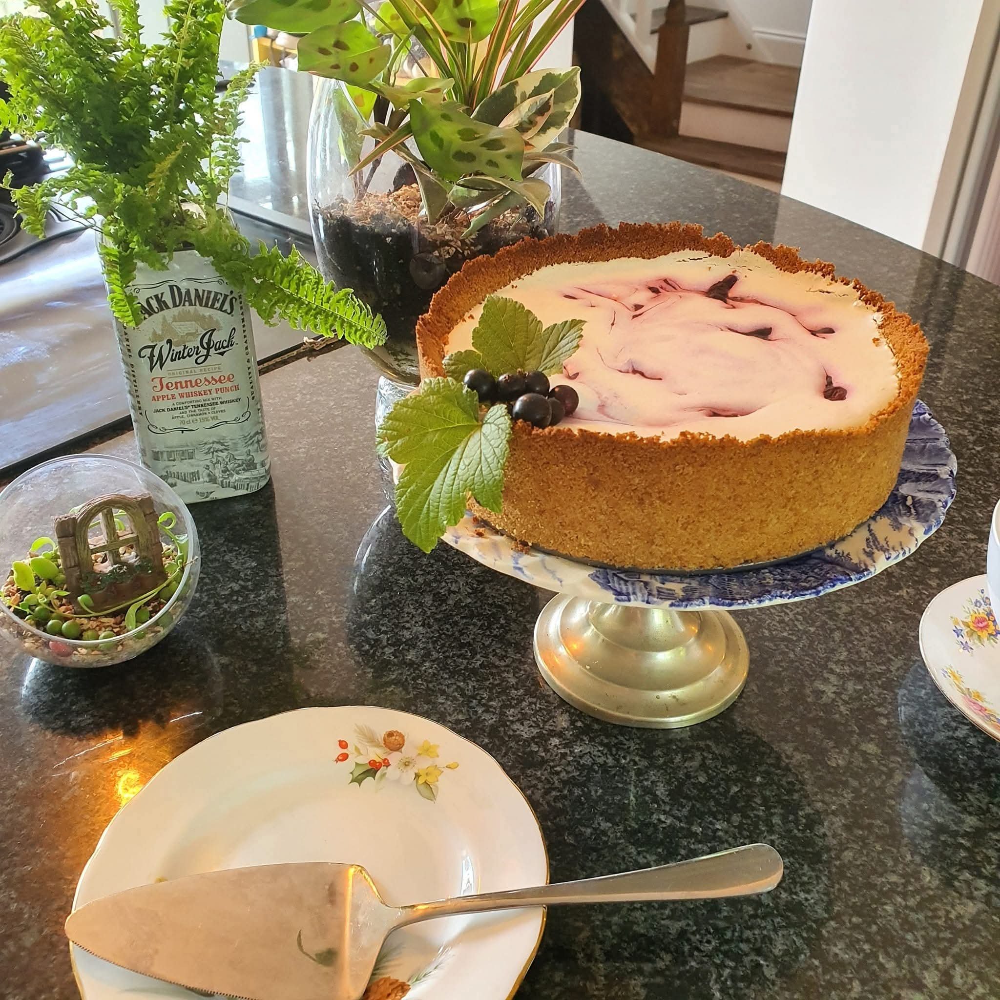 A close-up of a decorated birthday cake with pink marbled icing and a gold trim, placed on a blue and white patterned cake stand on a kitchen countertop. Surrounding the cake are decorative green leaves, a plant in a glass container, and a partially visible floral patterned plate.
