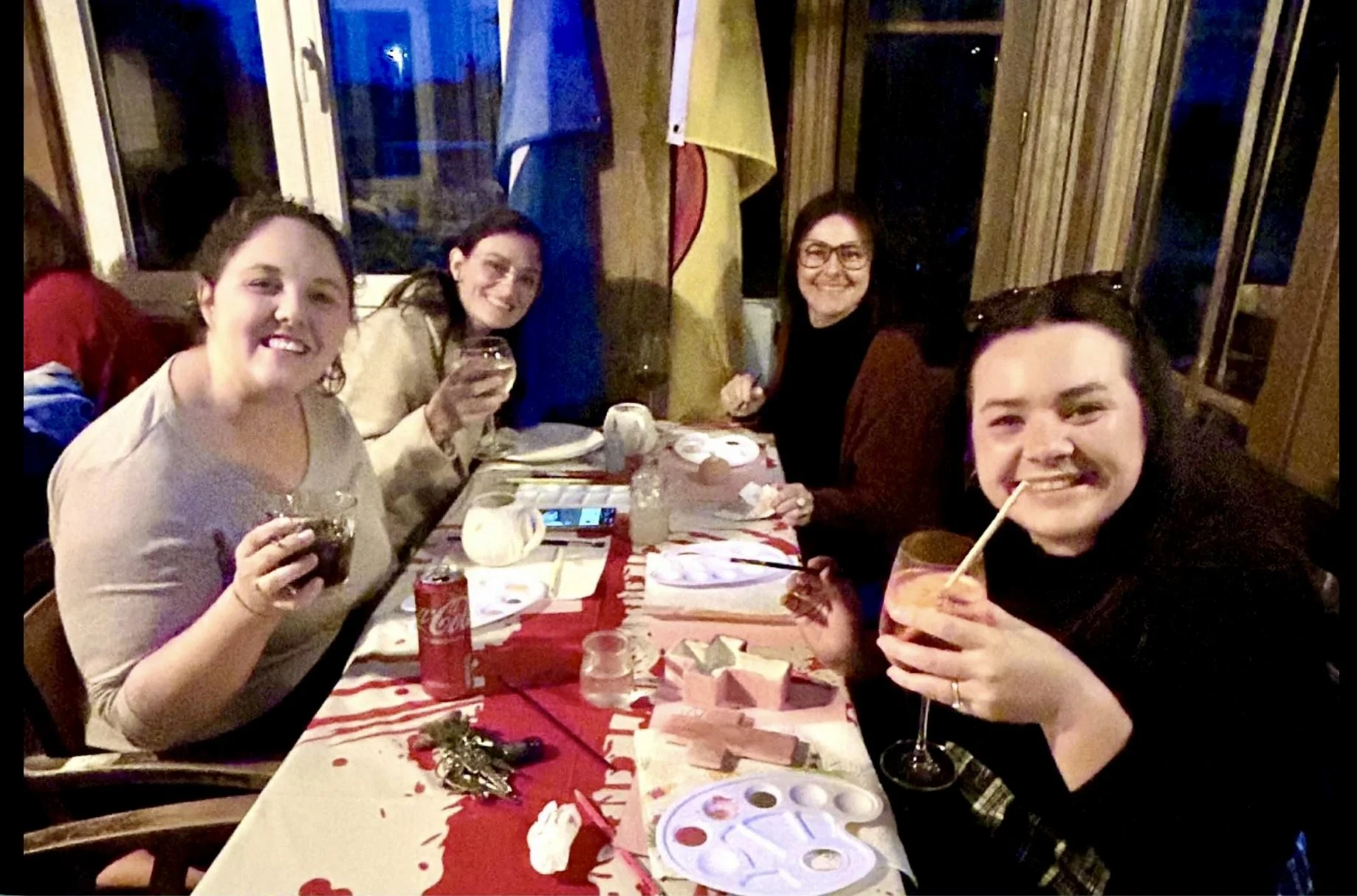 Group of five women sitting at a rectangular table with a red tablecloth, enjoying a celebration with drinks and desserts, in a cozy, warmly lit restaurant or home setting. They are smiling and looking at the camera.