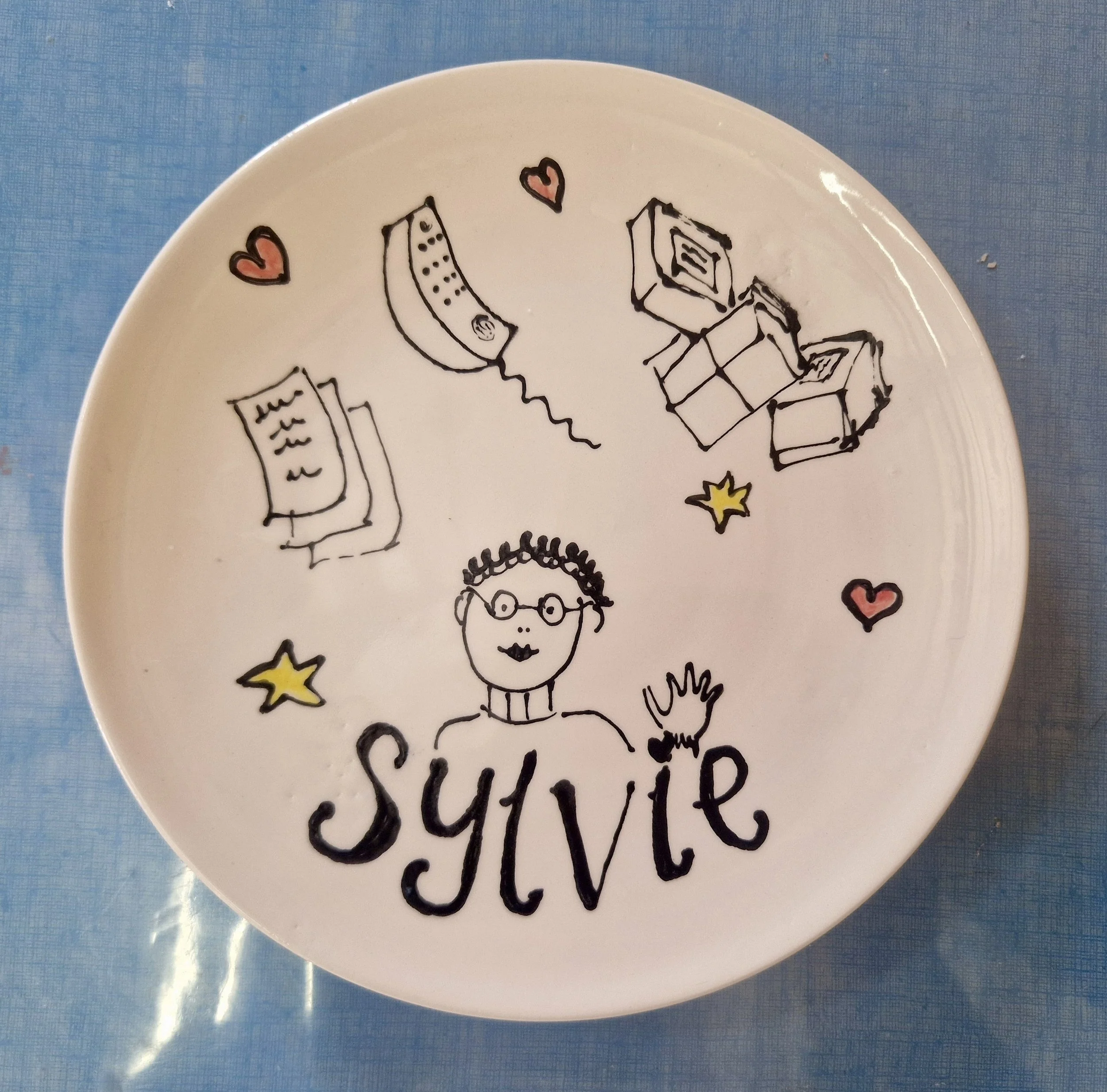 A white ceramic plate with black and red doodle drawings of a woman with glasses, a banana, a book, stars, hearts, and handwritten words, on a blue surface.