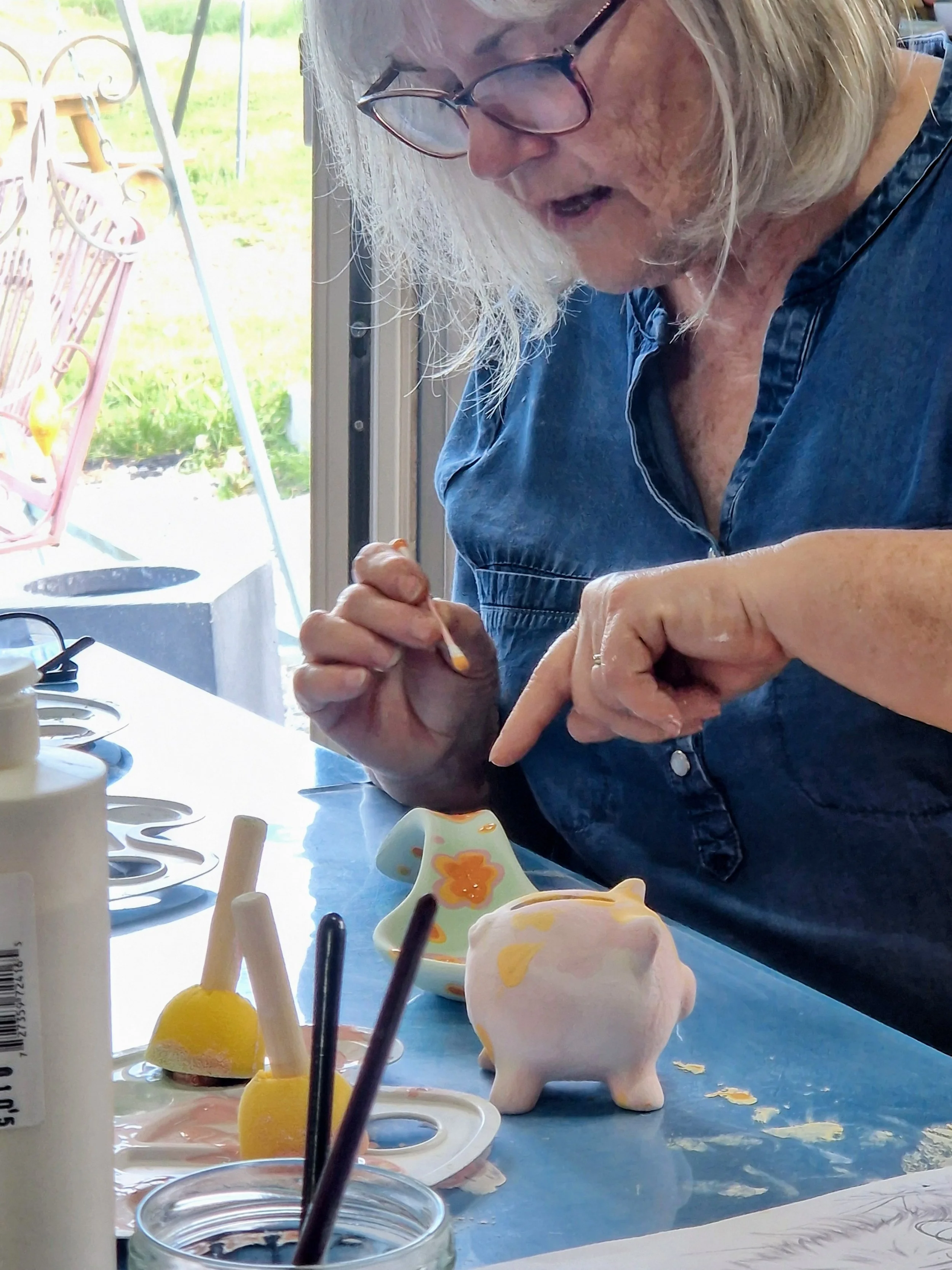 An elderly woman with glasses and blonde hair is decorating a pink ceramic piggy bank with yellow socks on a table filled with various colorful art supplies and ceramic figurines. She is wearing a blue denim shirt and working indoors near a window with a garden outside.