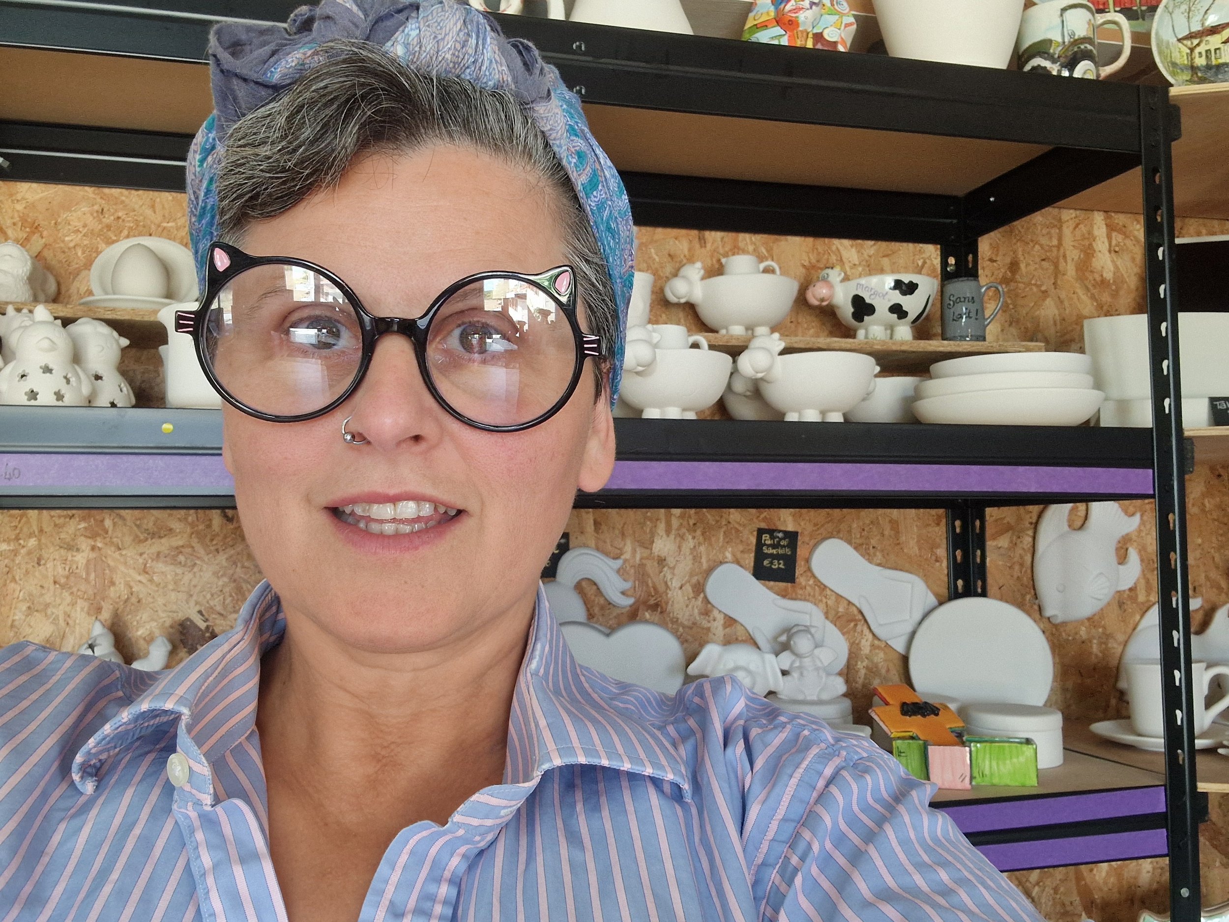 A woman with gray hair, wearing large, black, cat-eye glasses with pink accents, and a blue and purple patterned headscarf, smiling and taking a selfie in front of shelves filled with white ceramic dishes and cups.
