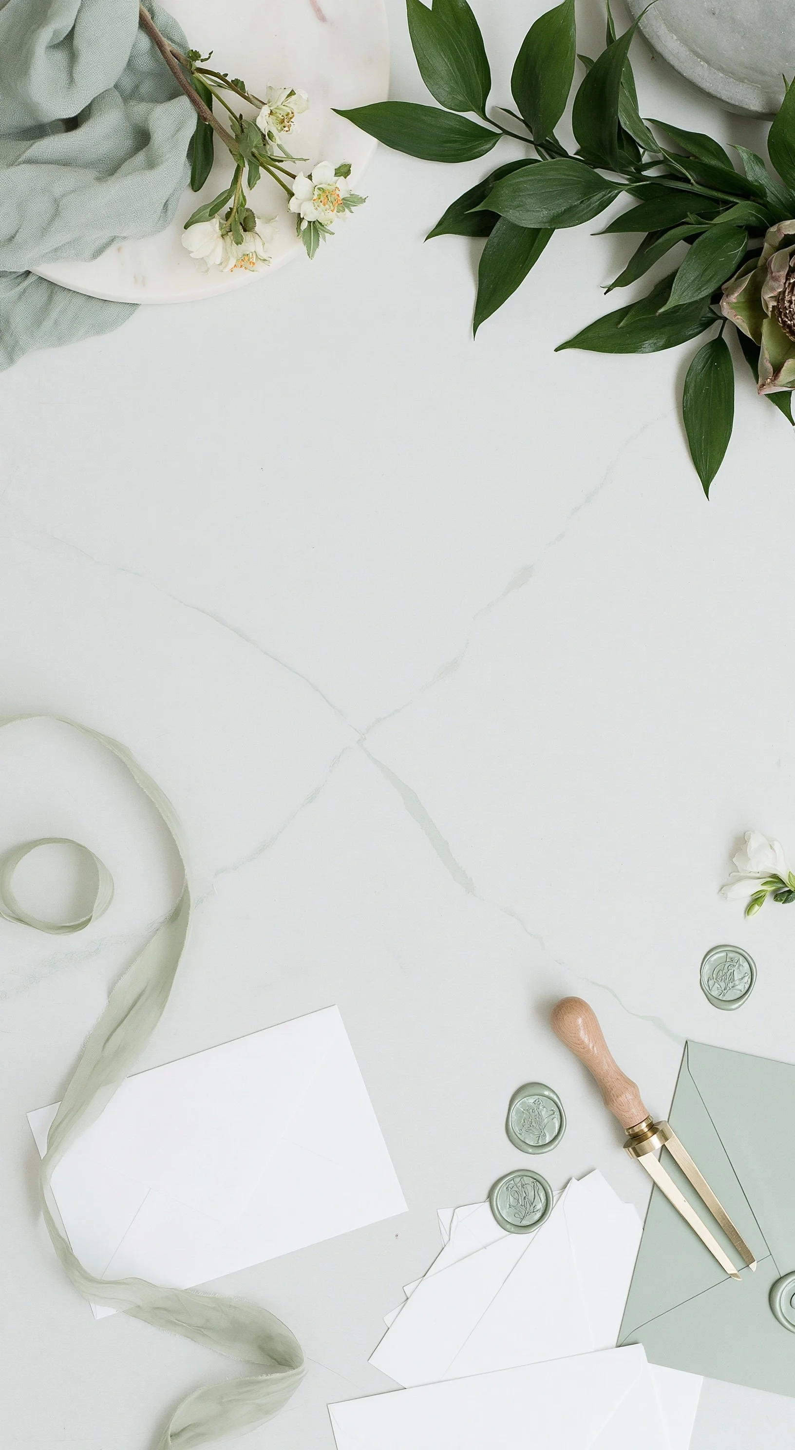 Flat lay of a clean white marble surface with stationery items including blank white envelopes, paper, a rubber stamp, wax seals, and a wax stamp tool. There are also green leaves and small flowers, creating a minimal and elegant aesthetic.