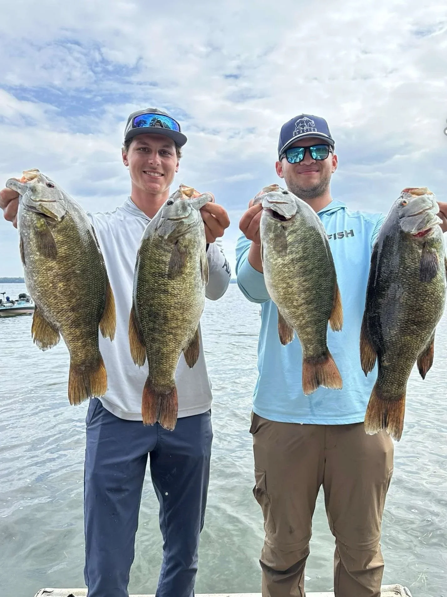 Two men standing in water, holding four large fish in front of them. They are wearing casual outdoor clothing and sunglasses, with cloudy sky above and boats in the background.