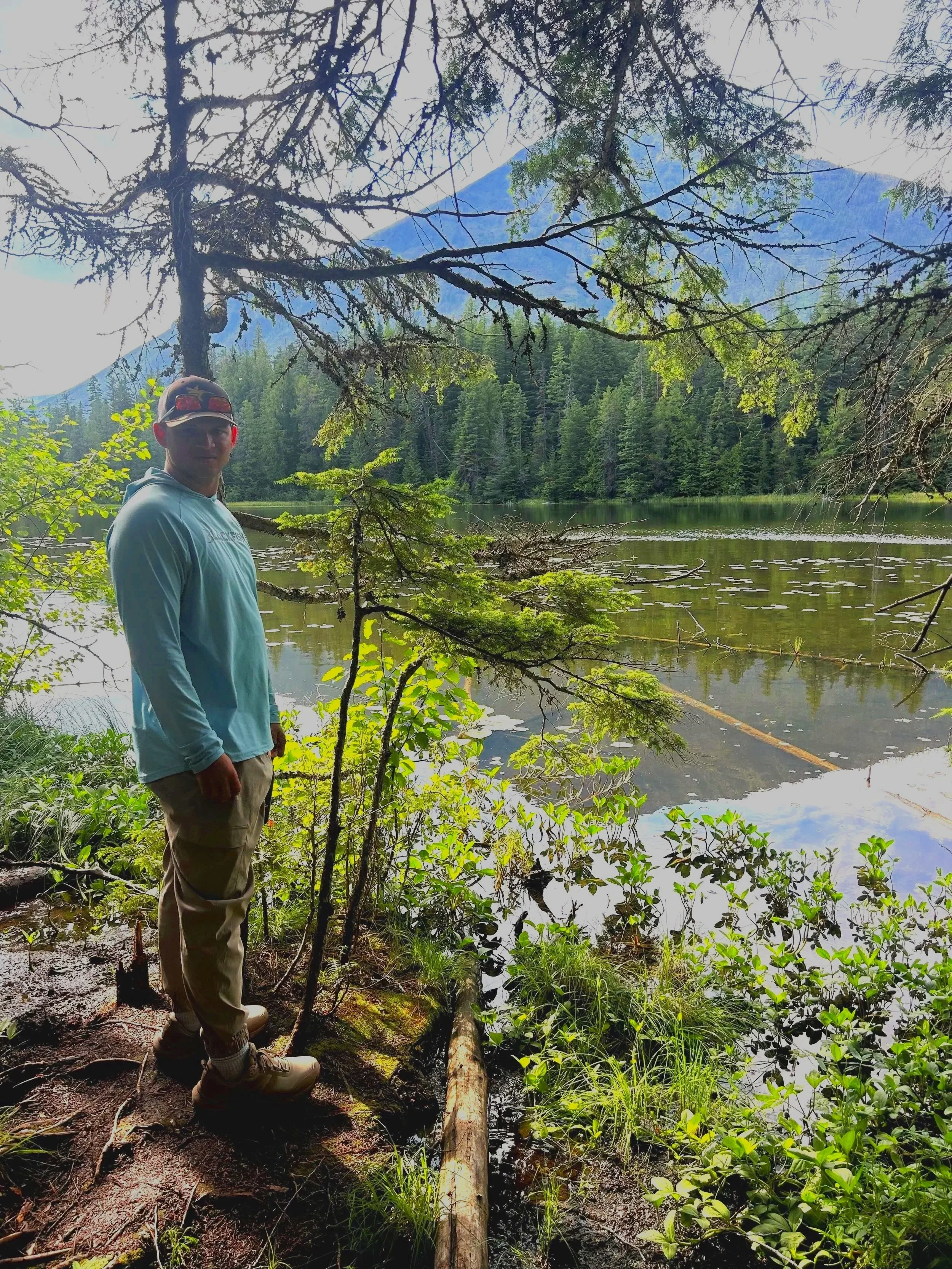 The Founder, Brandon Johnson standing near a lake surrounded by dense green forest with tall trees and mountains in the background, under a partly cloudy sky.