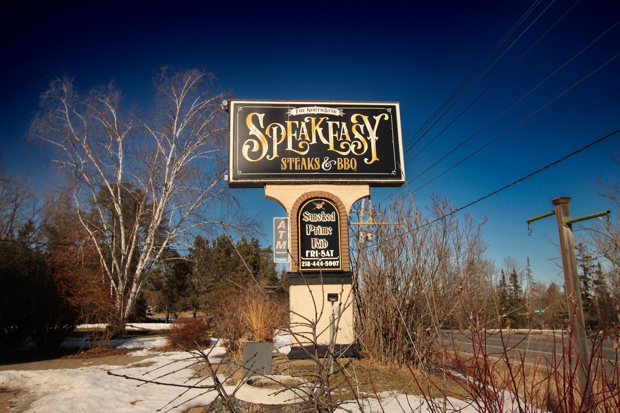 Sign for The North Star Steakhouse & BBQ with a menu on the lower part, set against a background of bare trees, a blue sky, and some snow on the ground.