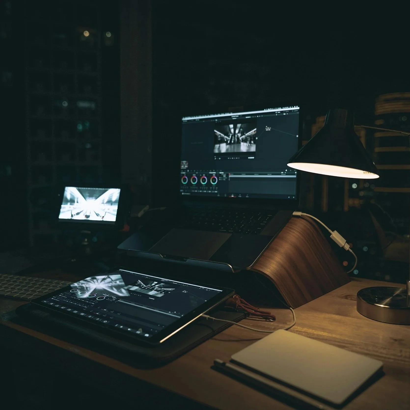 A workspace with a computer monitor, a tablet with a stylus, and a desk lamp in a dark room, showing a photo or video editing software and a black and white photo of an underground architectural space.