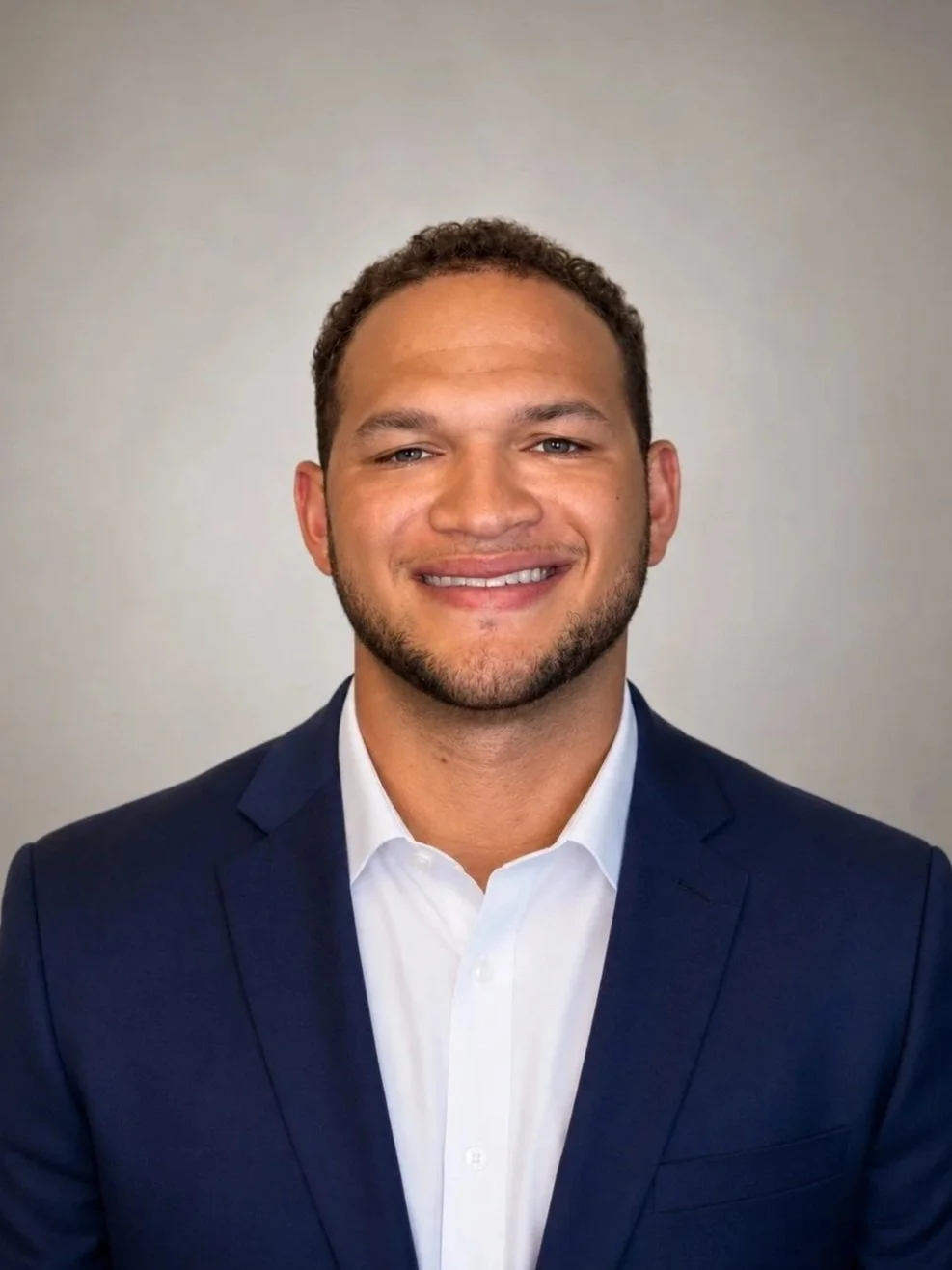 A young man in a navy suit smiling in a professional headshot against a plain background.