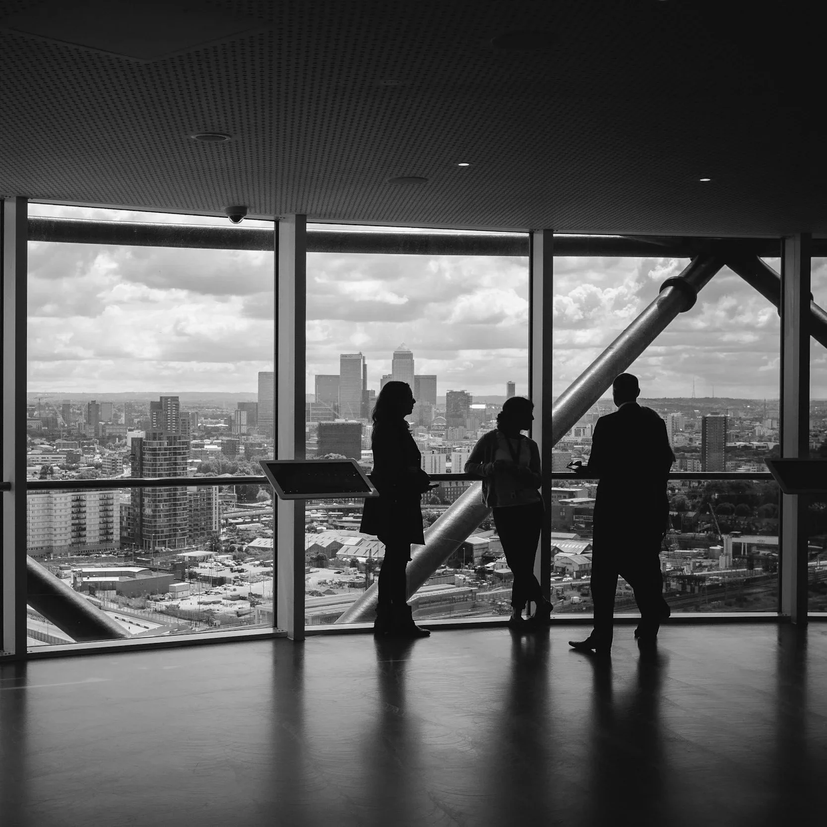 Silhouettes of three people standing and talking inside a high-rise building looking out at a city skyline through large windows during daytime.