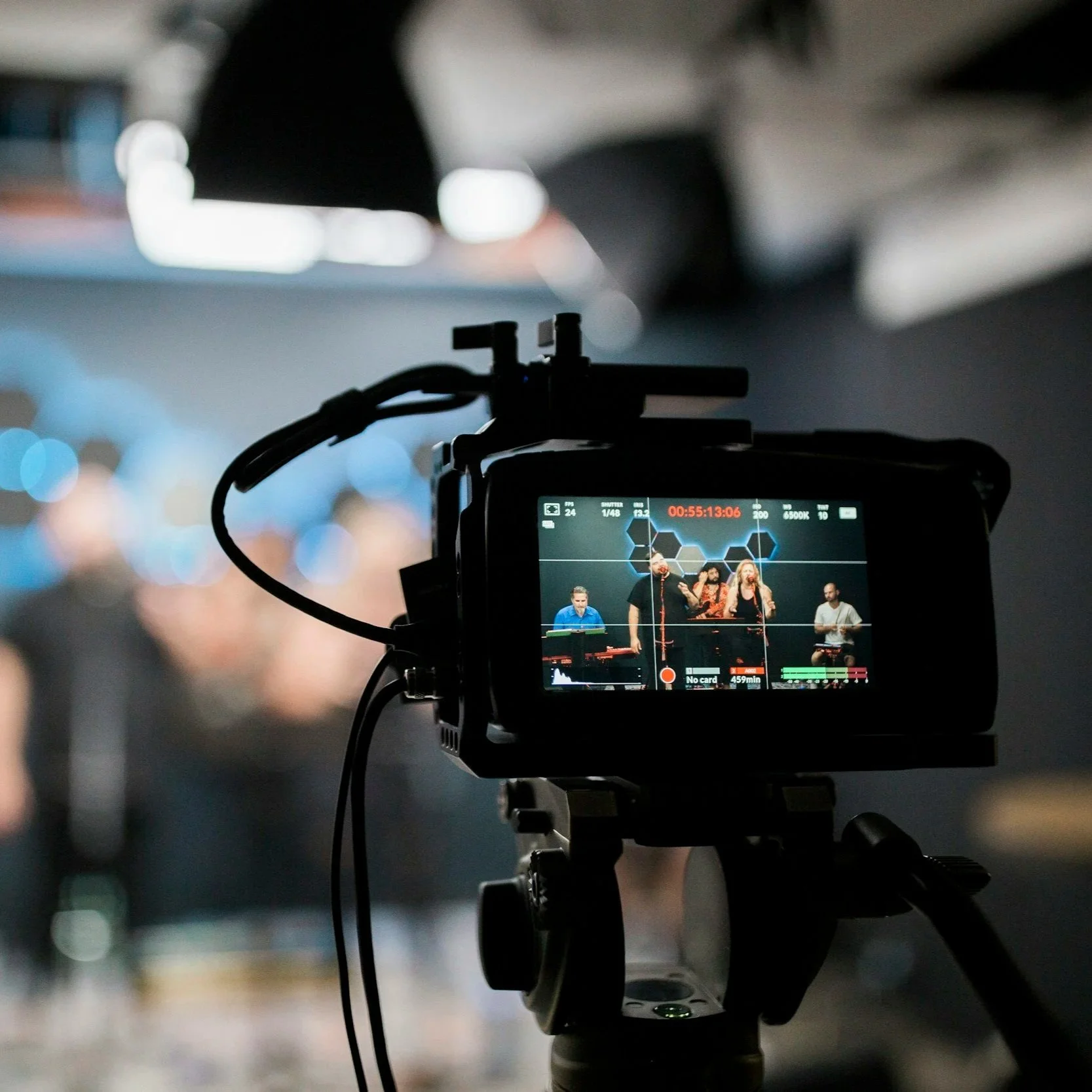 Photo of a video camera recording a band performing live on stage, with the camera's screen showing the band members and stage lighting.