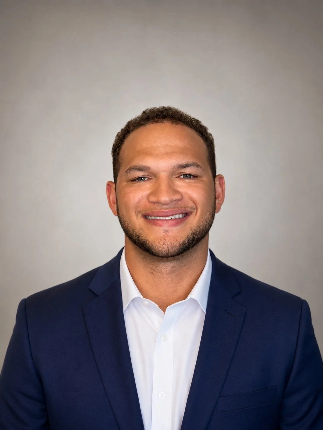 Headshot of a smiling man with short curly hair, wearing a navy blue suit and white dress shirt, against a neutral background.