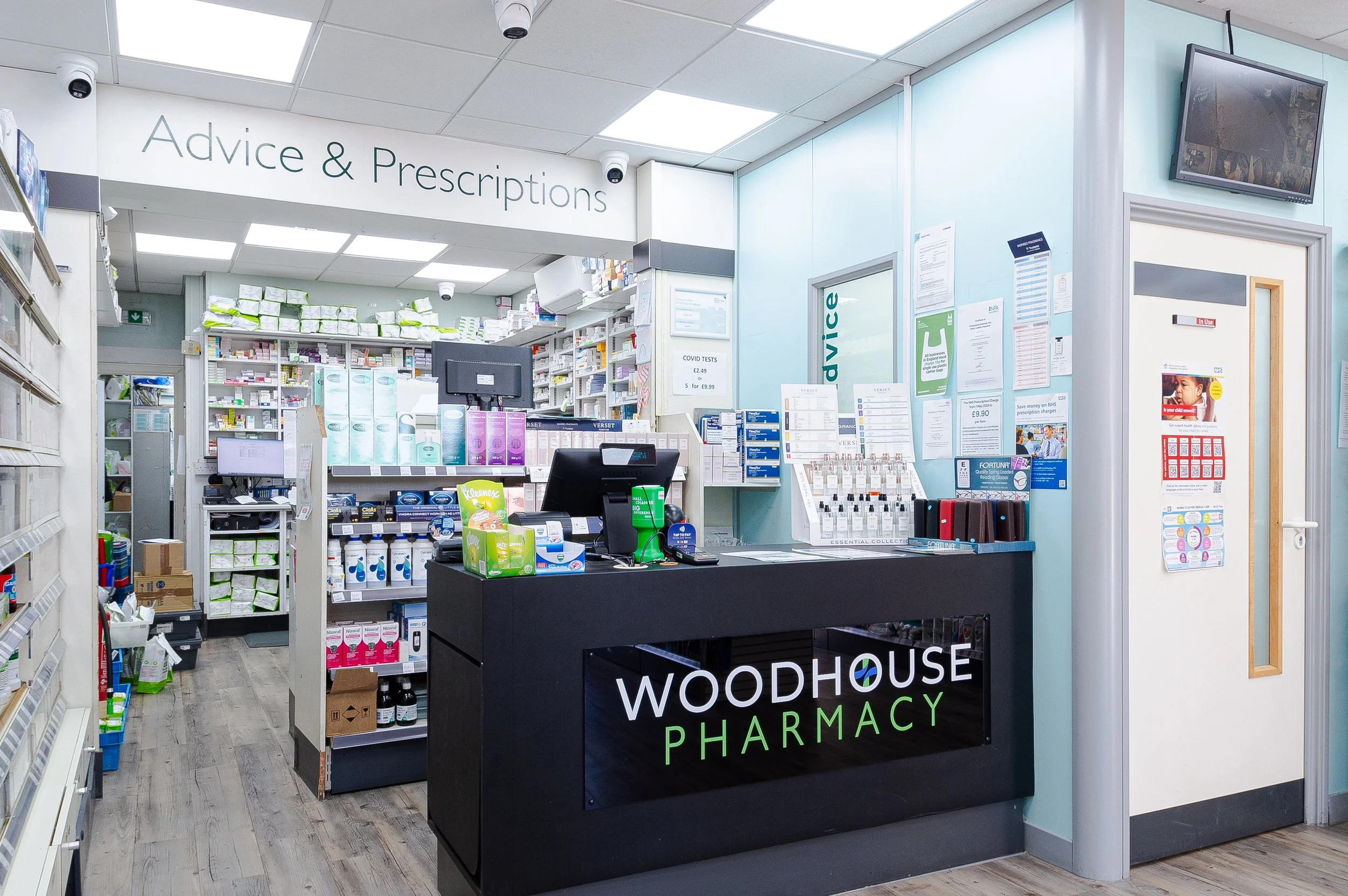 Interior view of a pharmacy, with a black counter labeled 'WOODHOUSE PHARMACY' at the front, shelves stocked with medicines and health products, a computer and cash register, as well as promotional posters and informational papers on the walls and gl