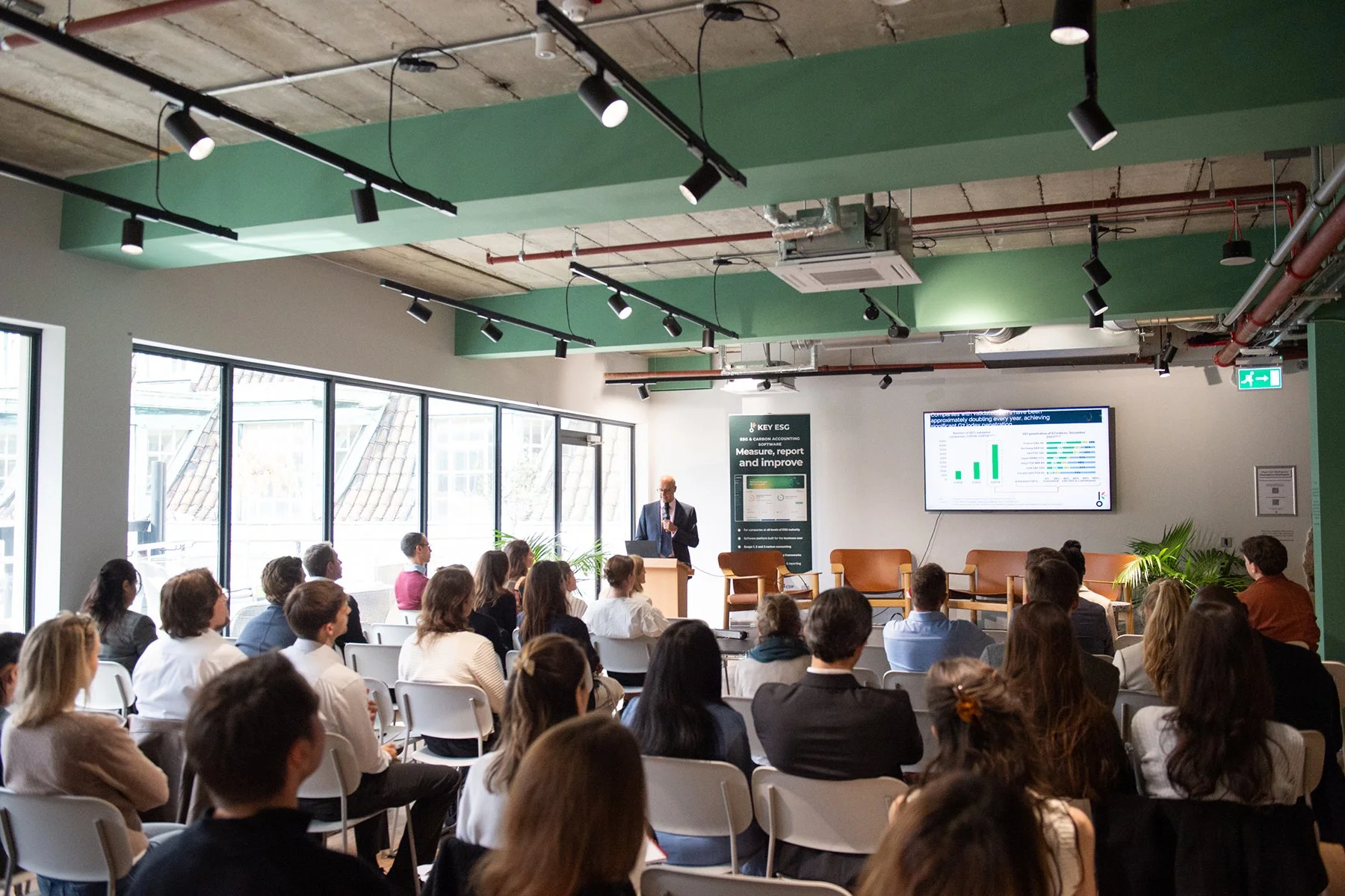 A presenter at a conference giving a speech to an audience in a modern, industrial-style room with large windows, green ceiling accents, and a screen displaying graphs and data.