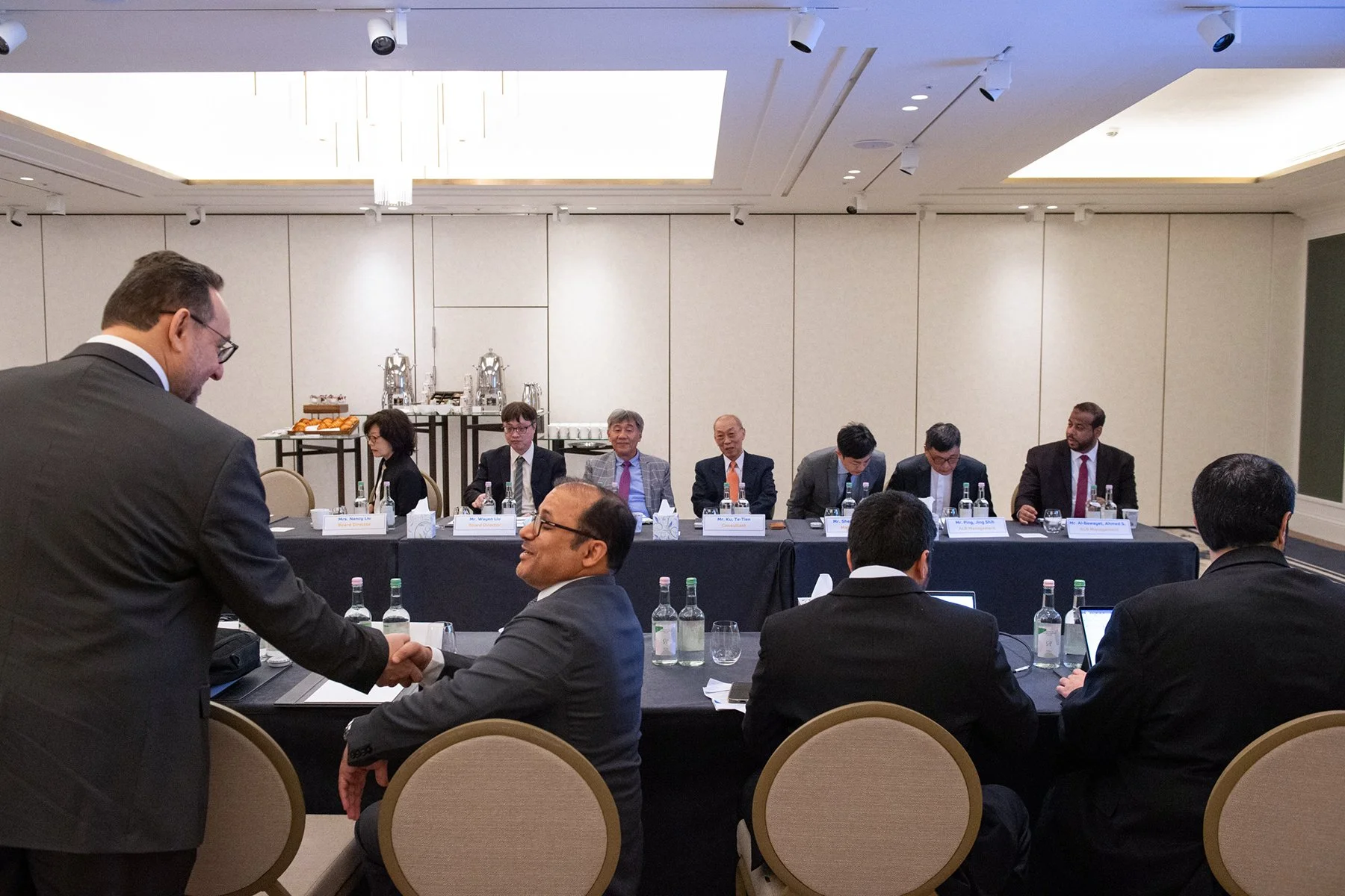 Business conference room with eight people seated at a long table covered with a black tablecloth. A man in a black suit is shaking hands with another man in a grey suit. Nameplates and bottled water are placed in front of each person. Two refreshmen