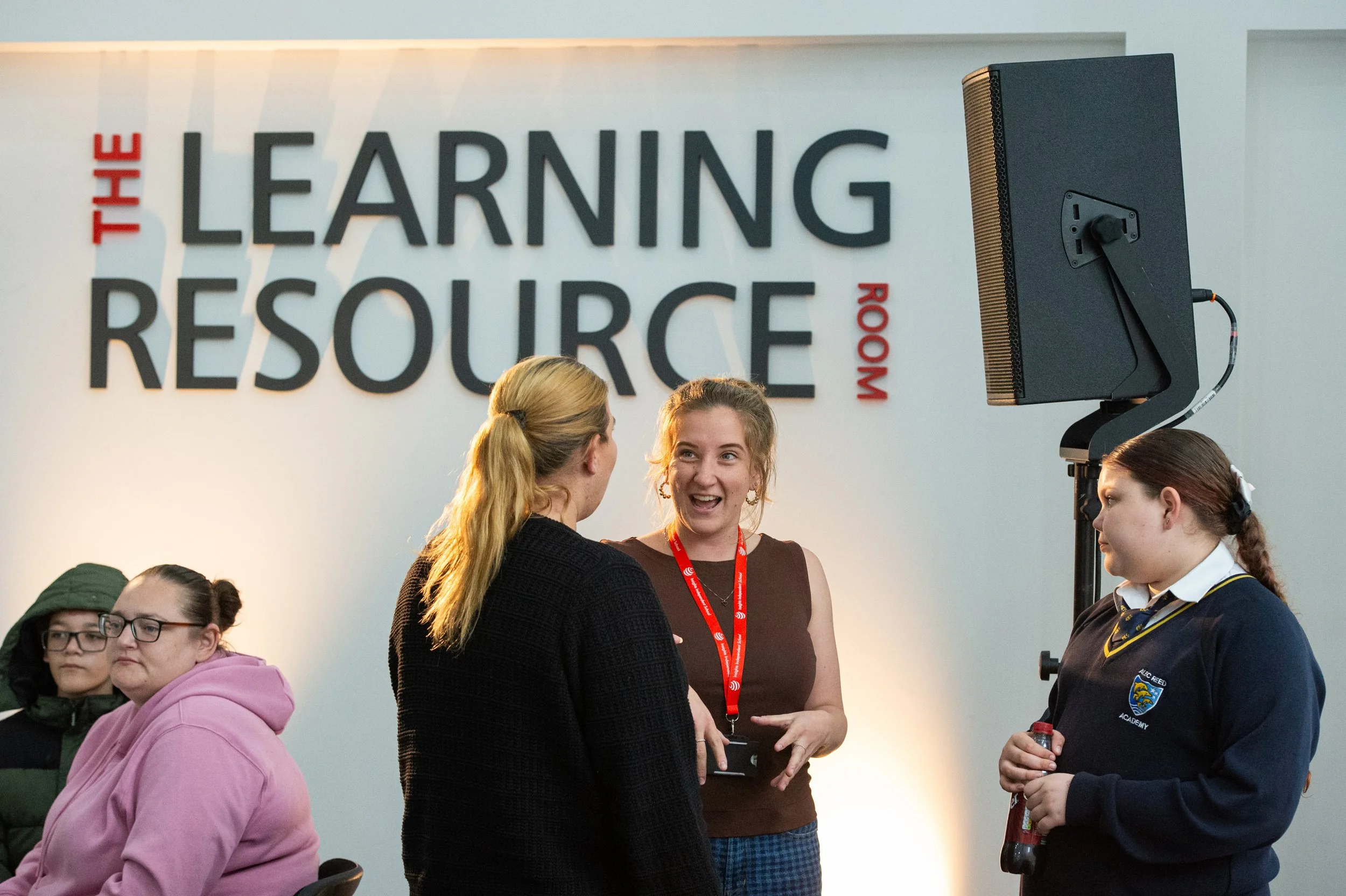 Group of people standing and talking at an indoor event with a sign that reads 'The Learning Resource Room' on the wall behind them.