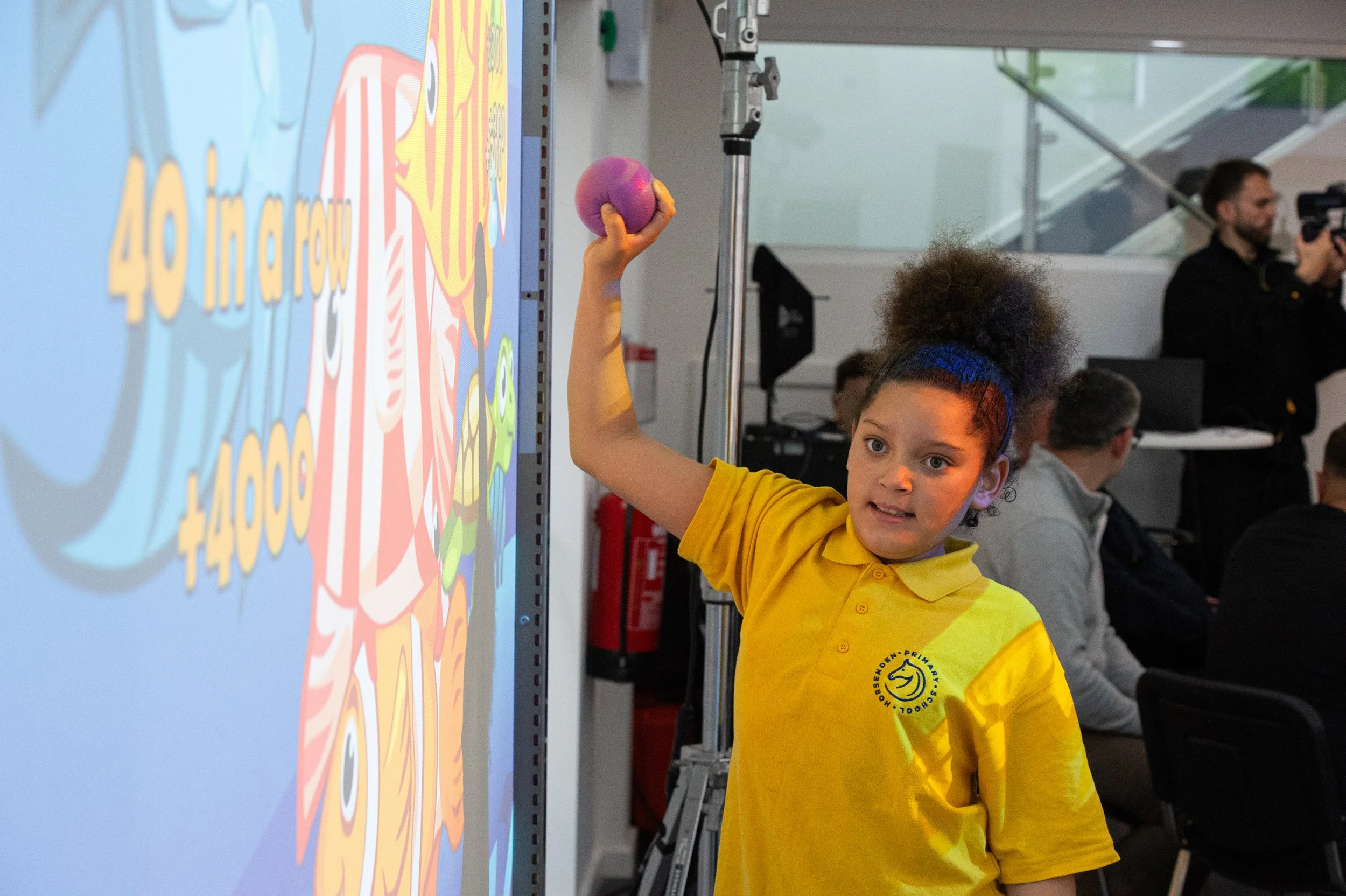 A young girl in a yellow shirt is raising her hand to hold a colorful ball, standing in front of a digital screen with vibrant graphics. Several people are seated in the background, with a man standing and holding a camera.