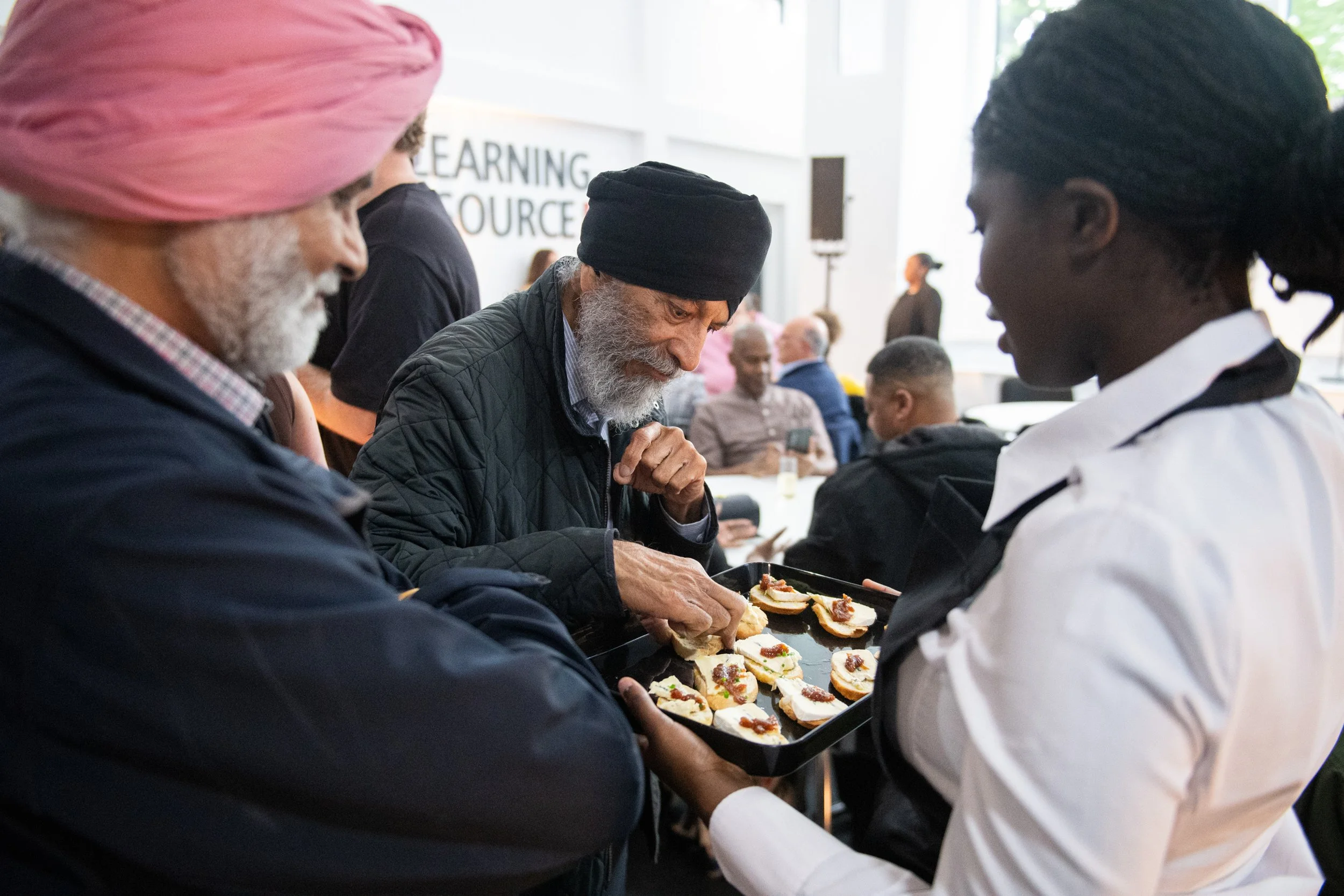 A diverse group of people gathered indoors, serving and eating appetizers, with a background sign that reads 'Learning Resources'.