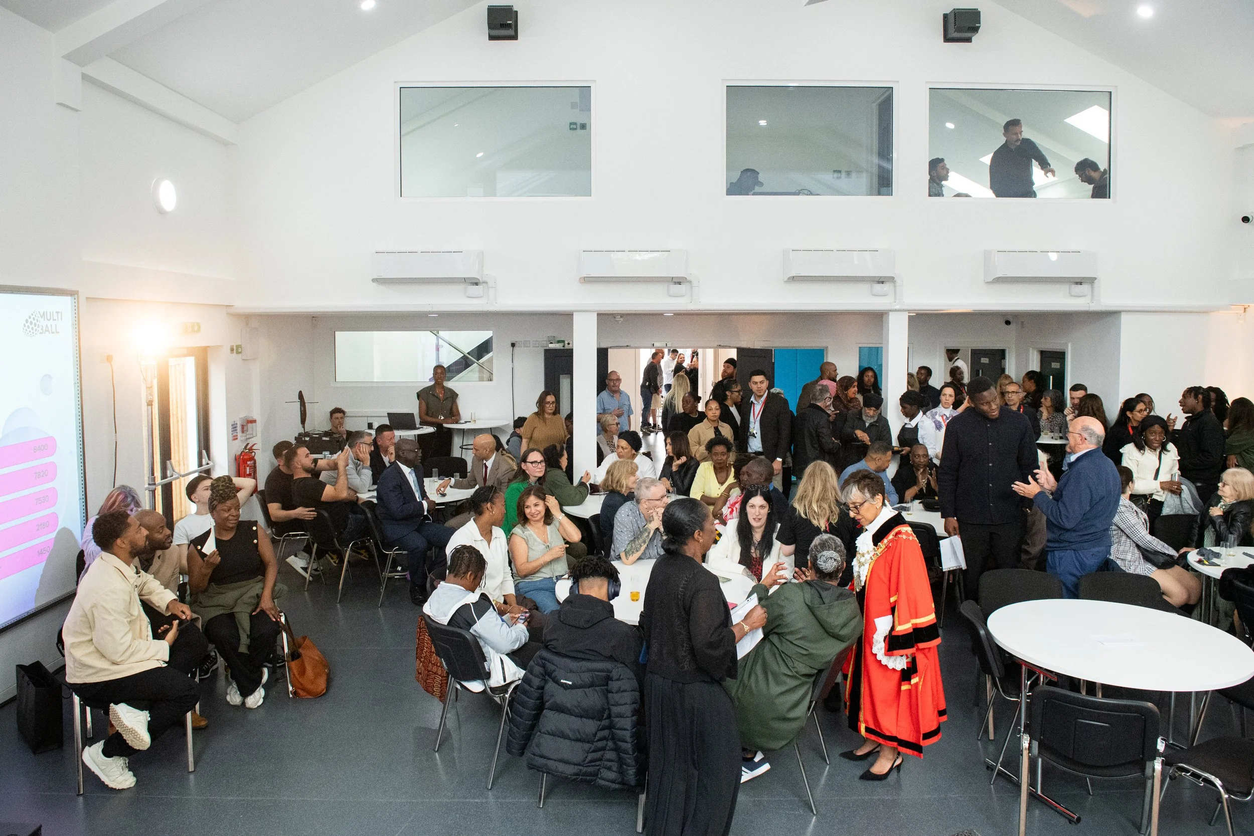 Large indoor event with many diverse people sitting at and standing around tables, engaged in conversations. There are windows, wall-mounted air conditioning units, and a stage area with a speaker in the background. The atmosphere appears lively and 