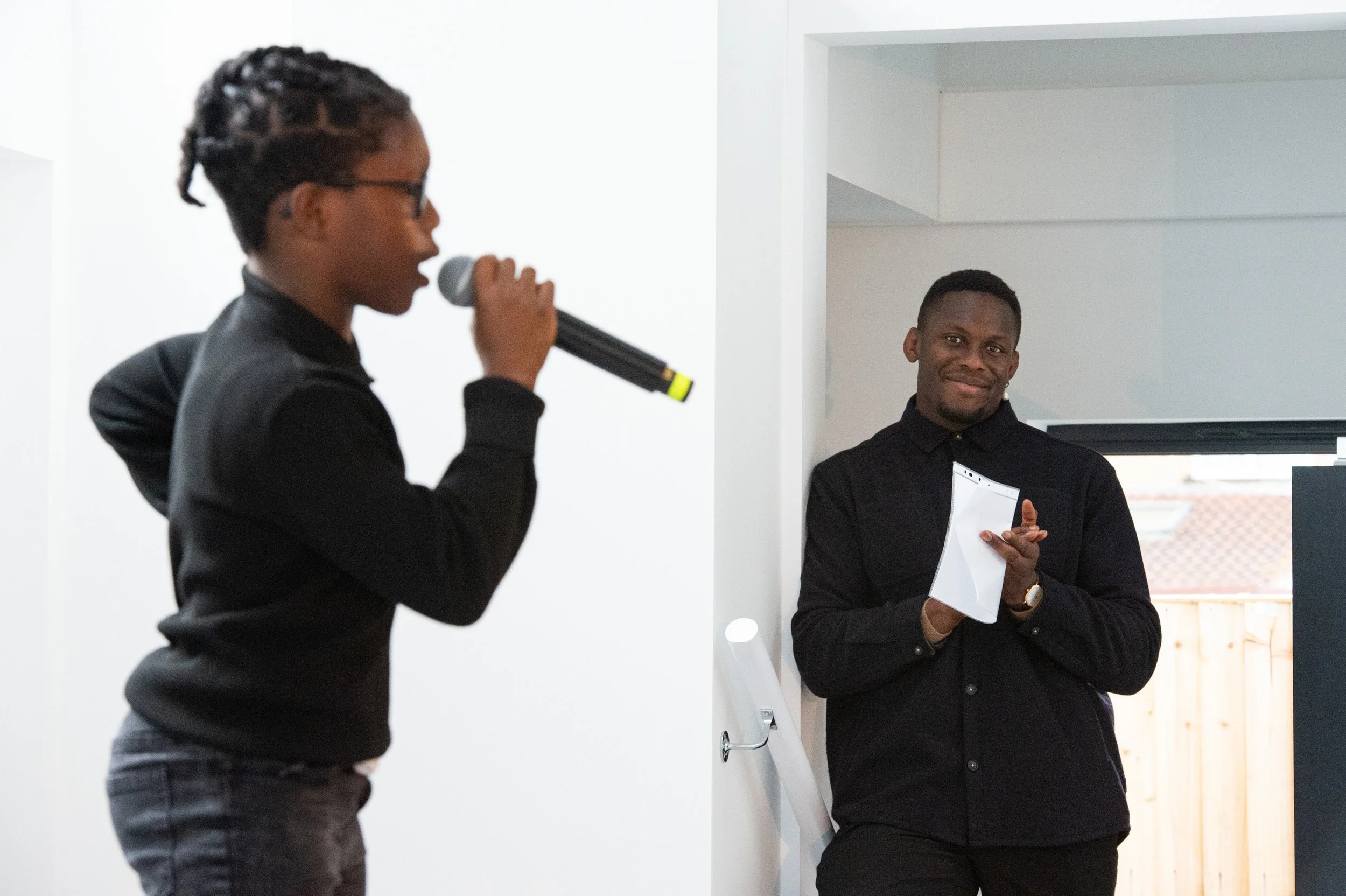 A woman with glasses, wearing a black jacket, is holding a microphone and speaking. A man, dressed in a black shirt, is standing nearby, leaning against a wall, holding a notebook, and smiling.
