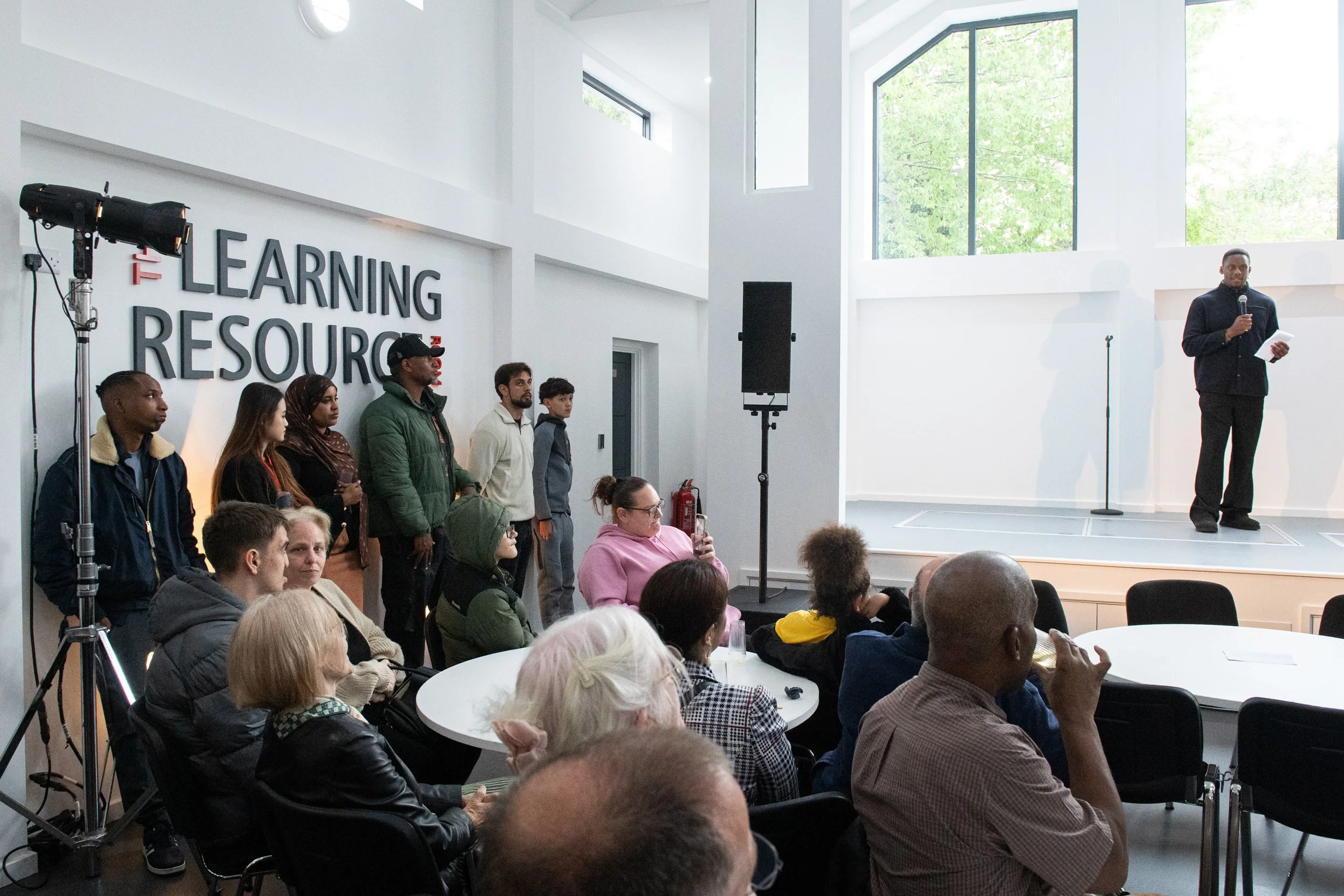 A speaker stands on stage with a microphone and paper in hand, addressing a diverse audience in a modern, bright room with large windows and a high ceiling. Some audience members are seated at round tables, while others are standing near a wall with 