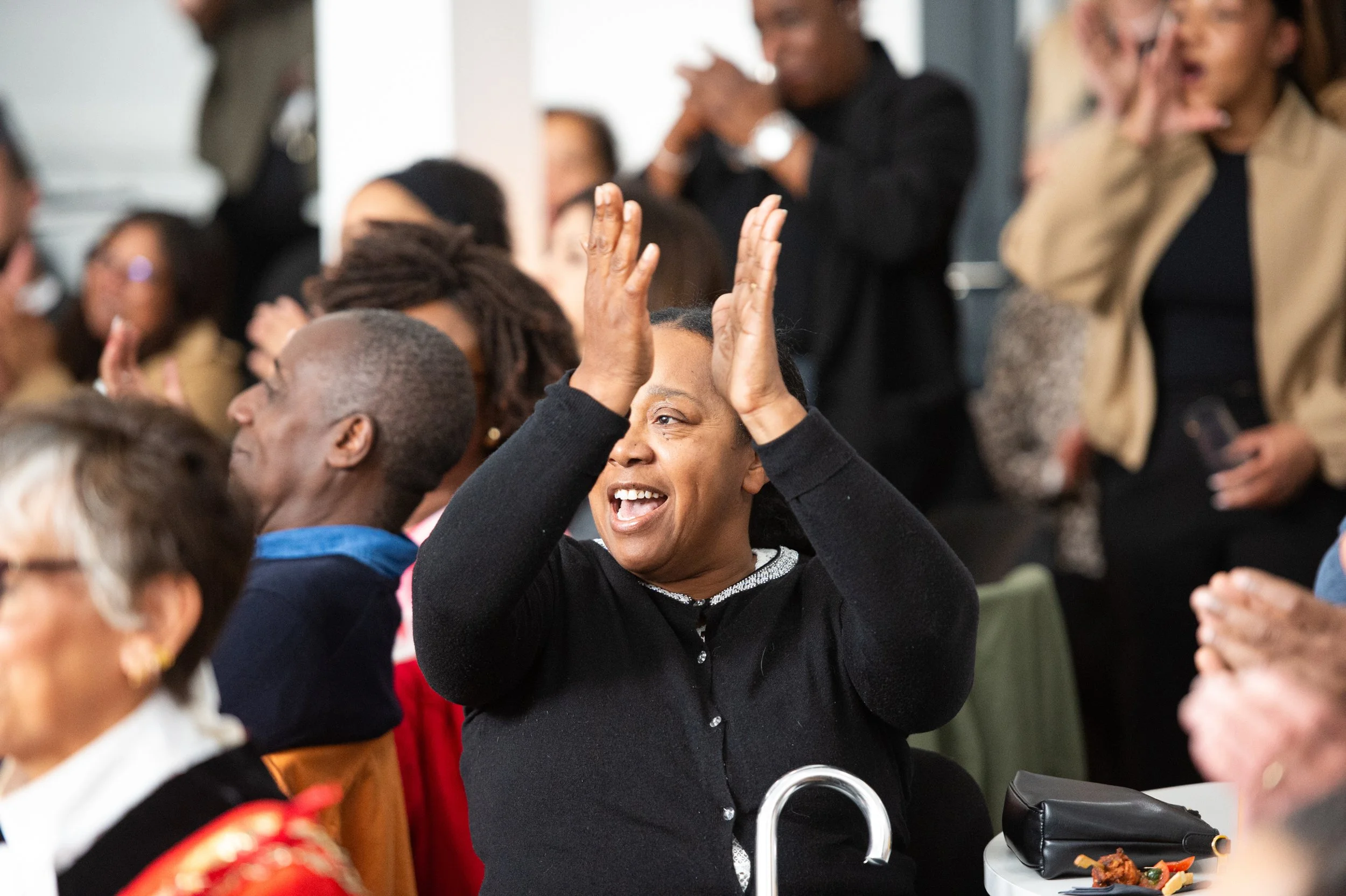 A woman with short black hair smiling and raising her hands in a celebratory gesture at a crowded event.