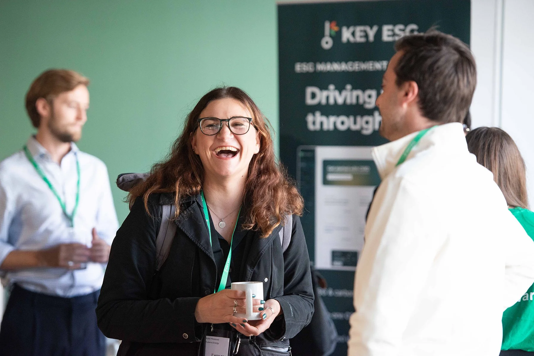Group of people at a conference, smiling and chatting, with a woman in the foreground holding a mug. A man in the background stands with a blurred badge, and a banner about ESG management is visible.