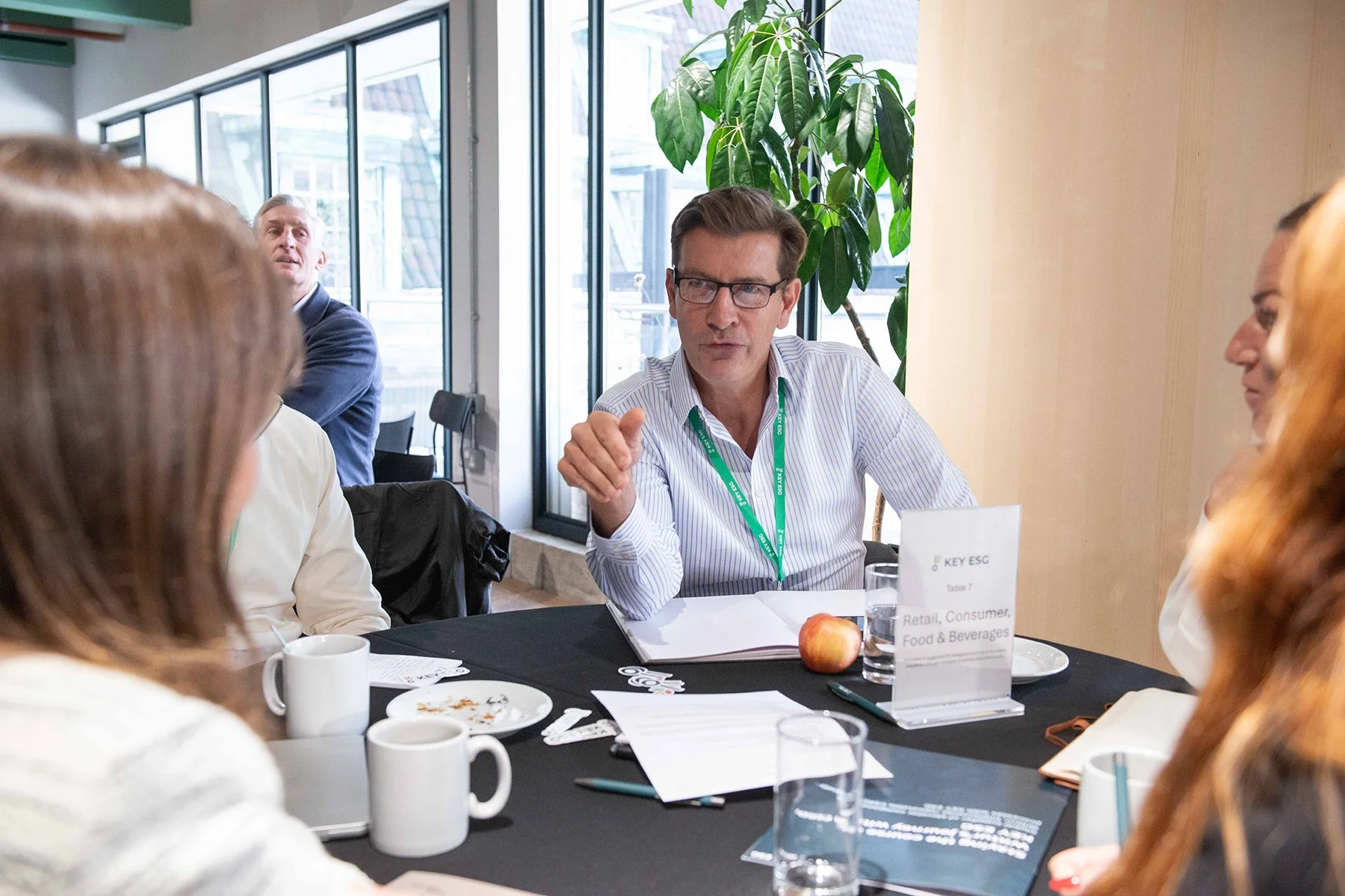 Group of people sitting around a table in a conference room, engaged in a discussion. The table has coffee cups, plates with leftover food, and a sign indicating they are part of a retail, consumer, food, and beverages discussion. A man wearing glass