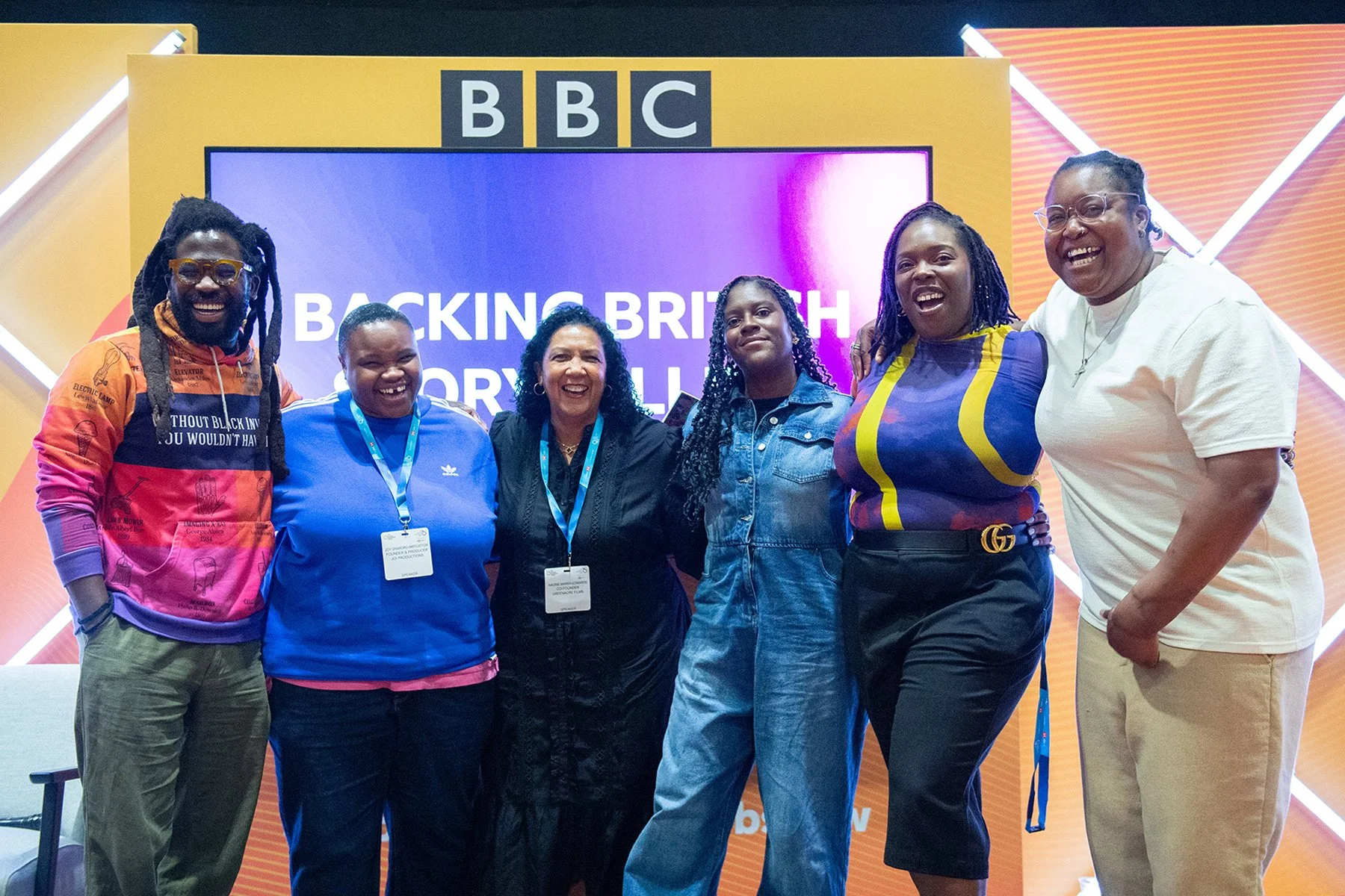 Group of six women standing together smiling at an event with a BBC backdrop, some wearing conference badges and lanyards.
