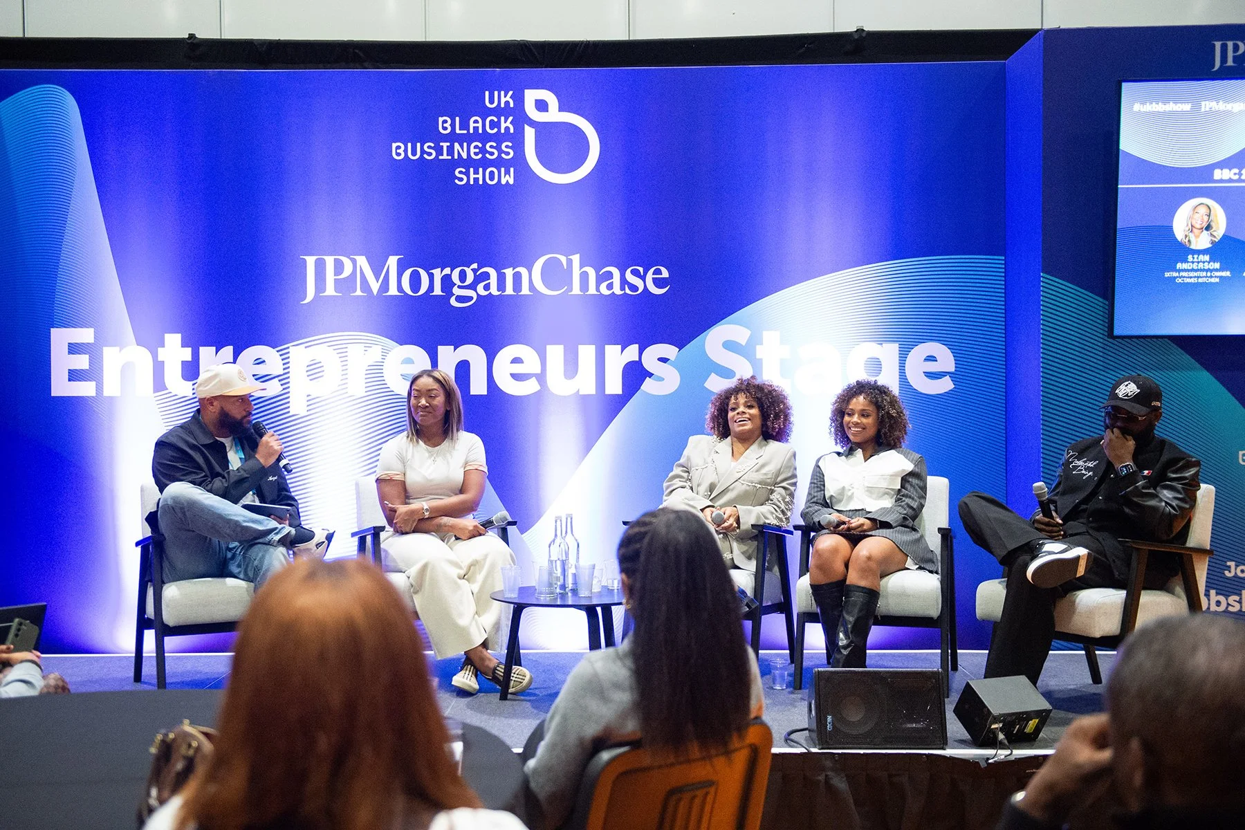 A panel discussion at the UK Black Business Show on the Entrepreneurs Stage, featuring five women and one man seated on stage with a blue backdrop displaying the event's logo and sponsors, including JP Morgan Chase.