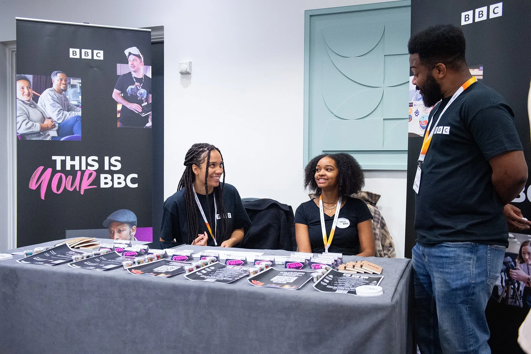 Two young women and one young girl sitting behind a table covered with black and pink promotional materials, chatting with a man standing in front of them. The background has banners with BBC logos and images of people, promoting the BBC. The women a