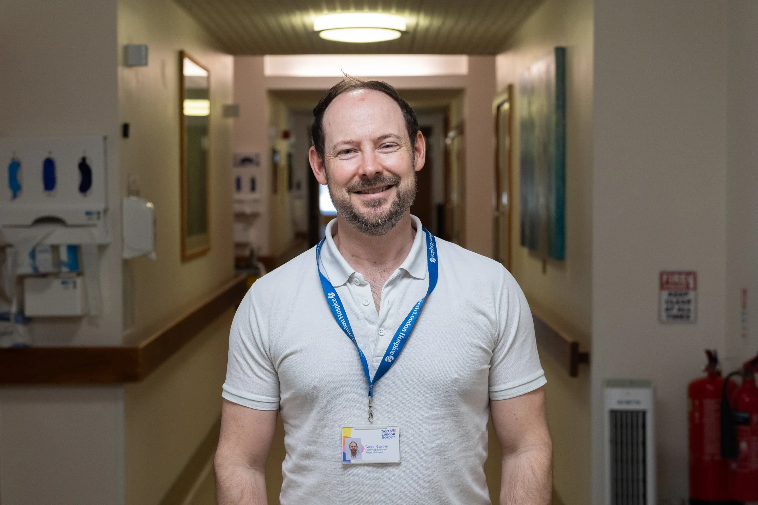 A smiling man with short brown hair and a beard, wearing a white polo shirt and a blue hospital staff badge, standing in a hospital hallway with artwork on the walls and medical equipment visible in the background.