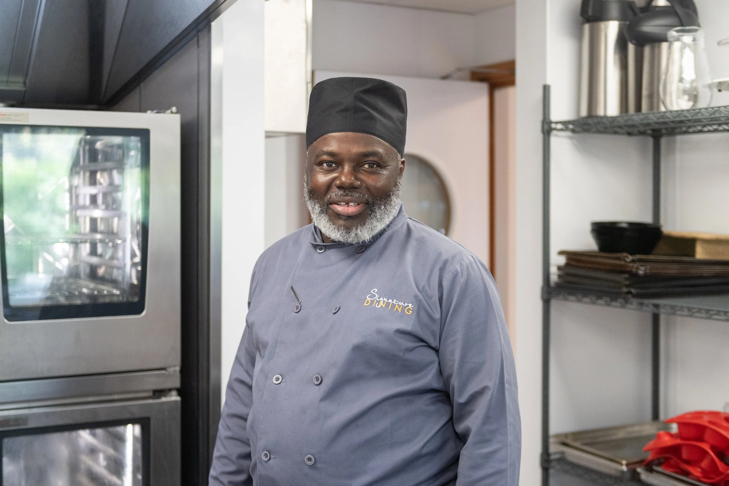 A smiling man in a gray chef's coat and black chef's hat, standing in a commercial kitchen with stainless steel appliances and racks of baking trays in the background.