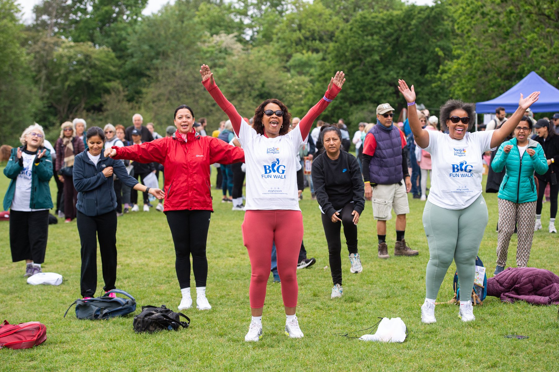 Group of diverse people participating in an outdoor walking event, some with arms raised, on a grassy field with trees and a blue tent in the background.