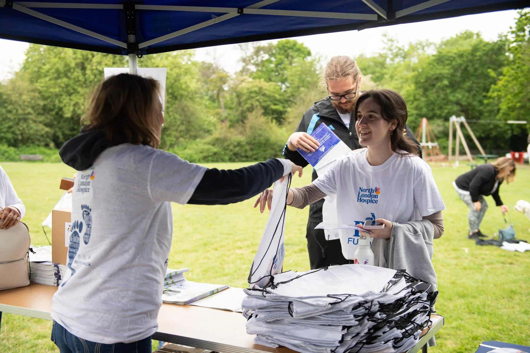 People participating in an outdoor event under a canopy in a park, with stacks of t-shirts and informational materials on a table, and children playing in the background.