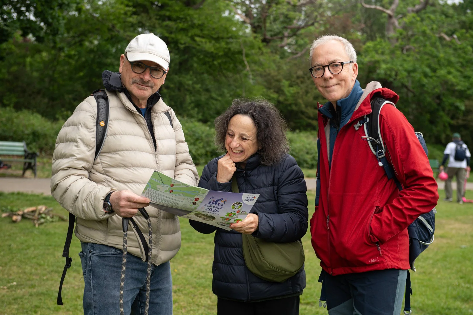 Three adults, two men and one woman, are outdoors in a park with green trees in the background. They are wearing jackets and backpacks, and they are looking at a colorful map together, smiling.