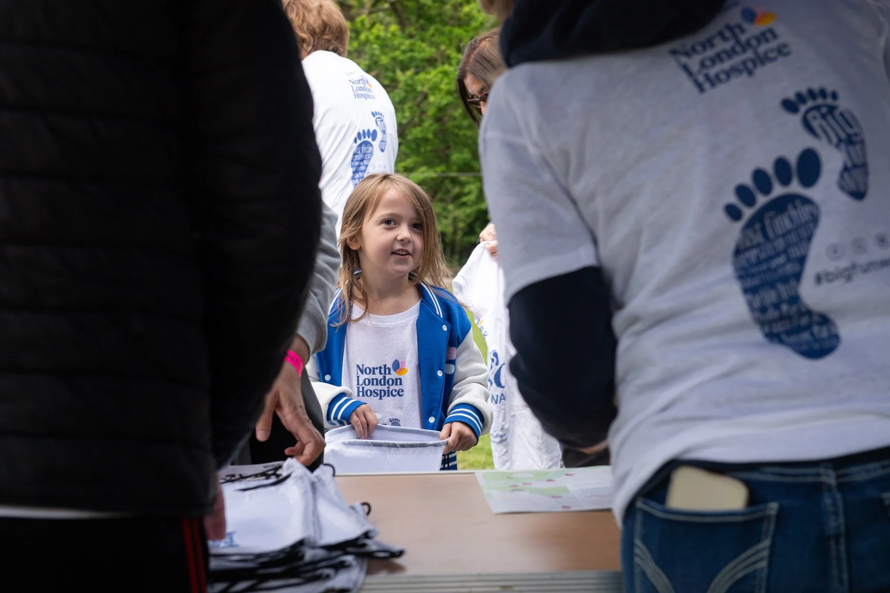 A young girl is participating in a charity event for North London Hospice, surrounded by volunteers wearing white T-shirts with the hospice's logo. The girl is holding a container and is engaged in the activity outdoors with trees in the background.