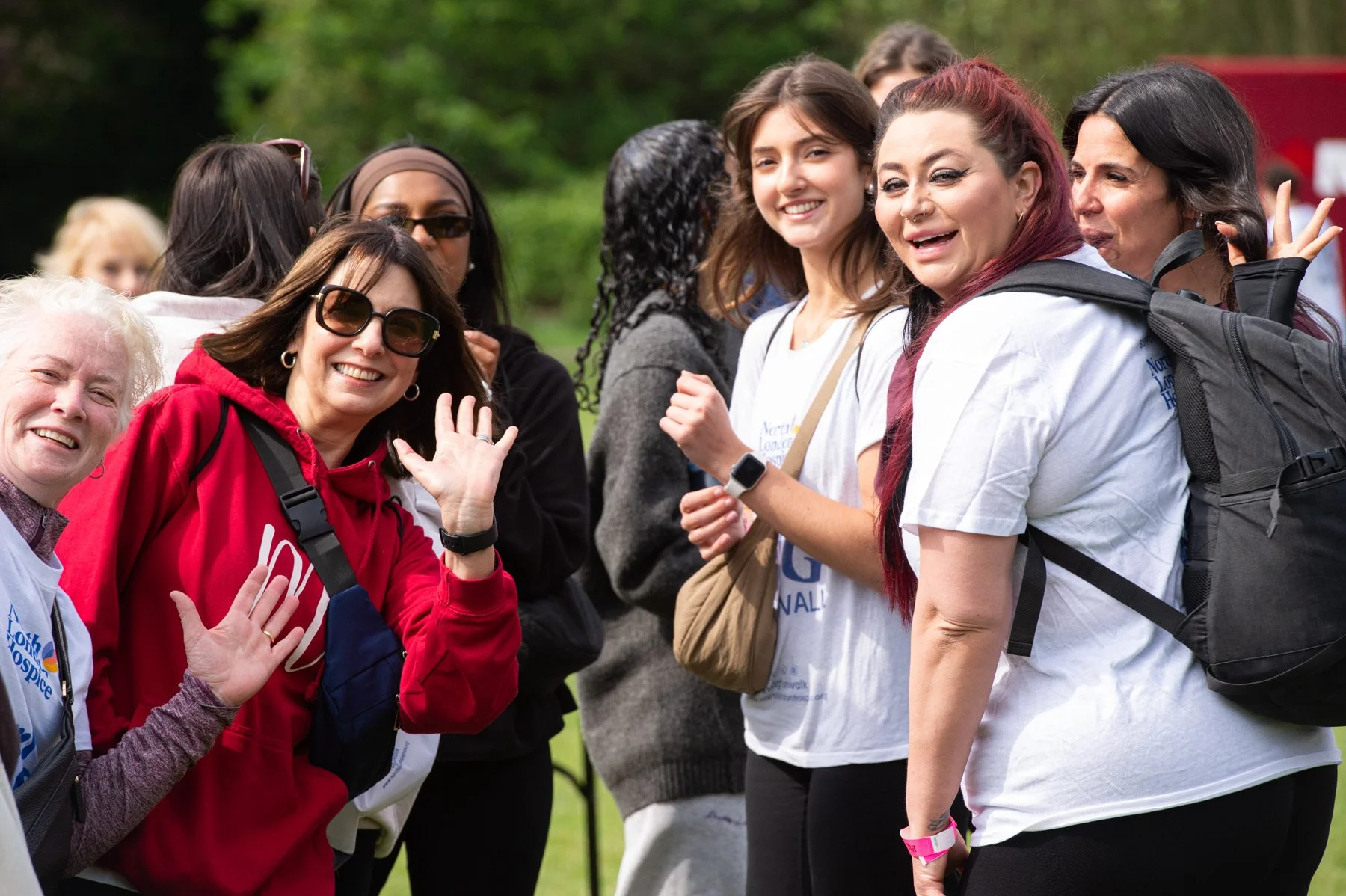 Group of smiling women gathered outdoors, some waving at the camera, with trees in the background, participating in a charity event or walk, wearing casual clothing and event badges.