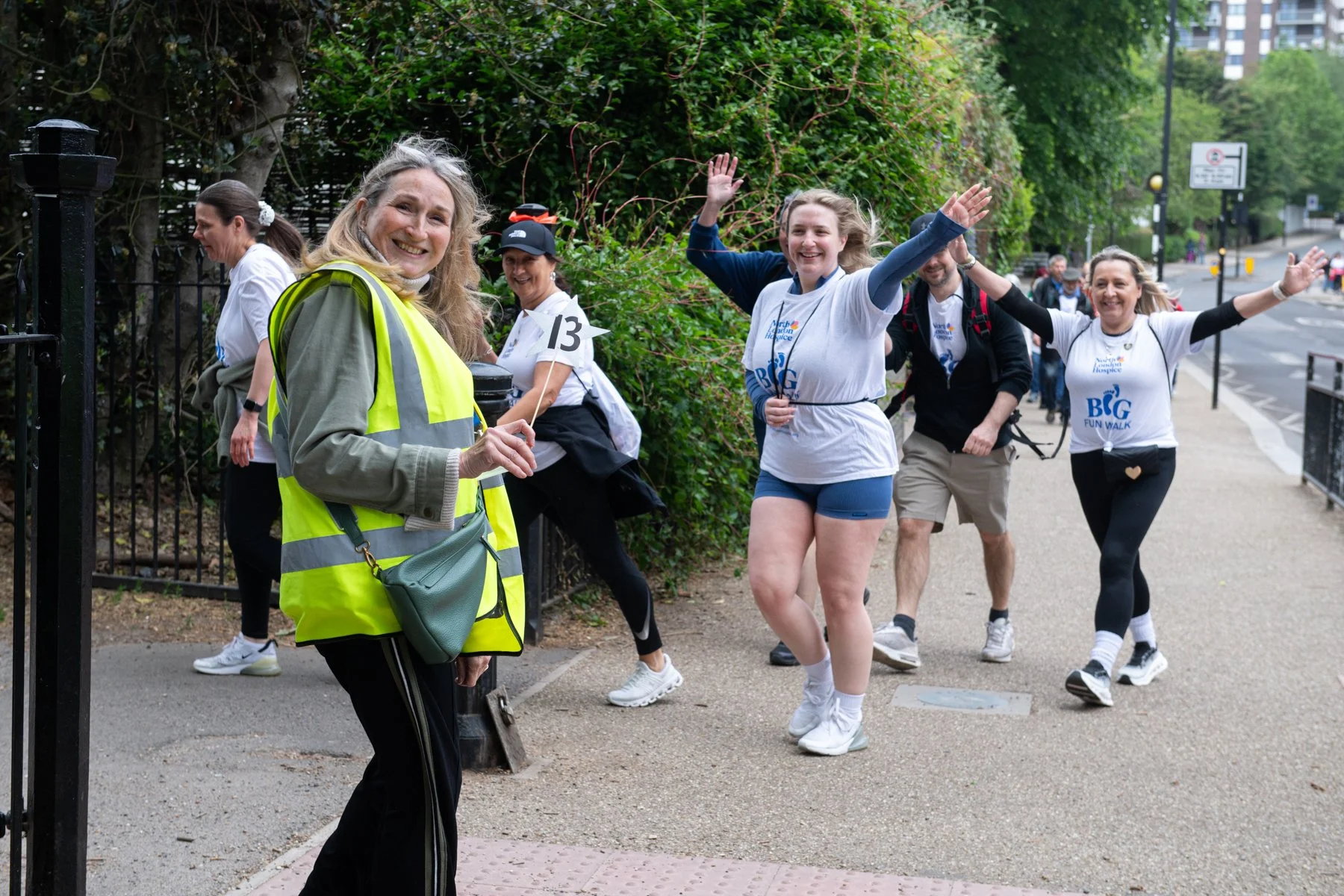 Women participating in a charity walk event, wearing matching white t-shirts with the event name "Big Fun Walk," some with backpacks, walking along a sidewalk on a cloudy day, with a volunteer woman at the front holding a small flag and smiling.