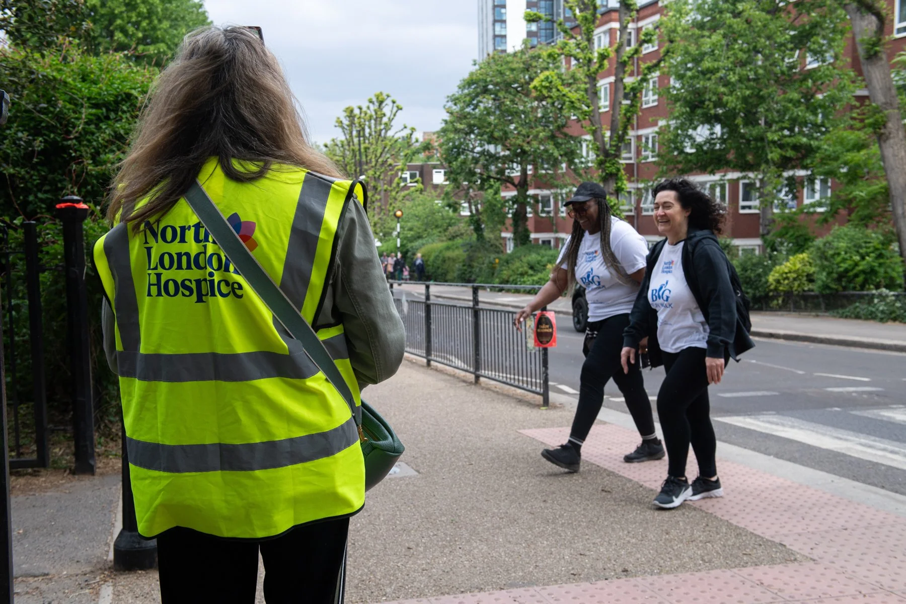 A person wearing a yellow vest with the text 'North London Hospice' stands on a sidewalk, facing two women crossing the street and smiling. The women are laughing, and there are trees and residential buildings in the background.