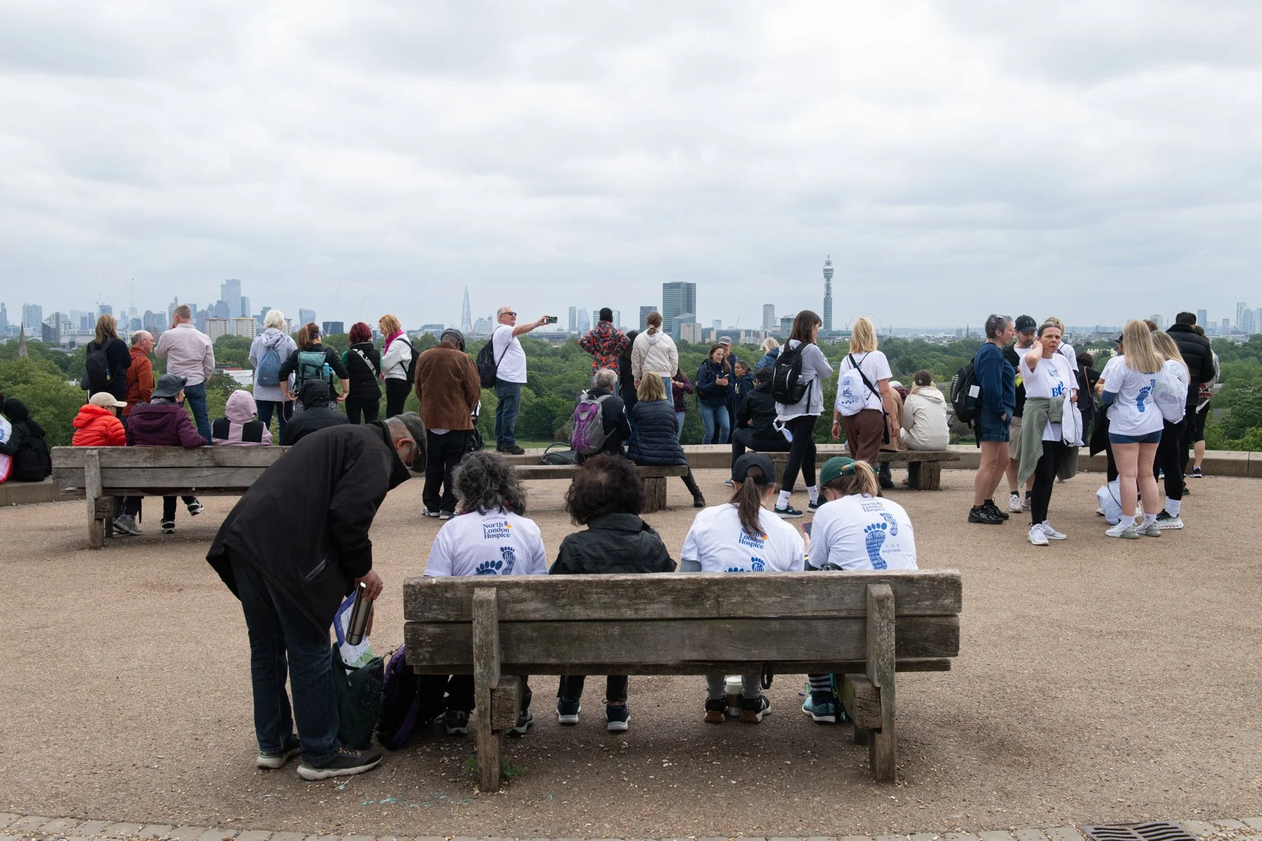 A diverse group of people gathered outdoors on a cloudy day, some sitting on benches and others standing, with a city skyline featuring tall buildings and a tower in the background.