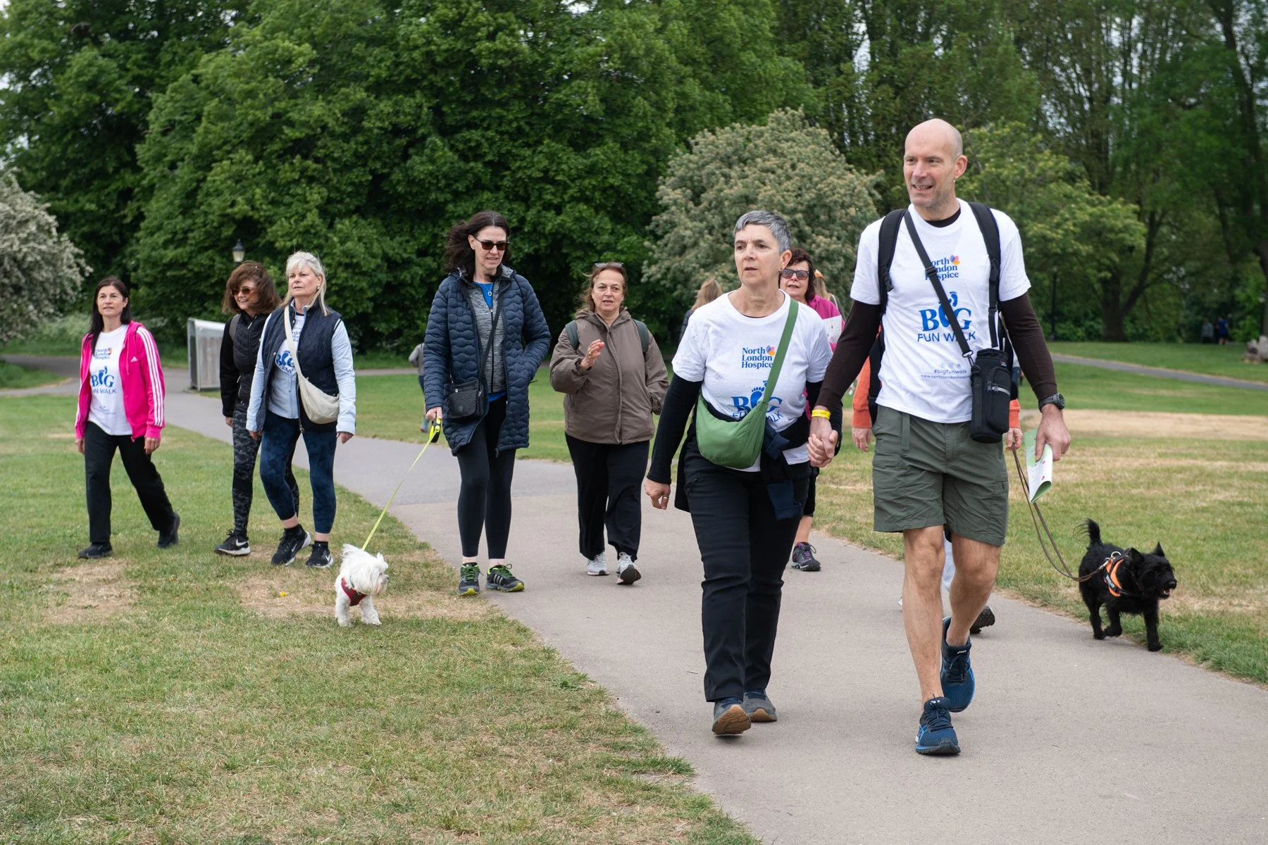 A group of people participating in a charity walk in a park, wearing event t-shirts, walking dogs, and enjoying a cloudy day with trees in the background.
