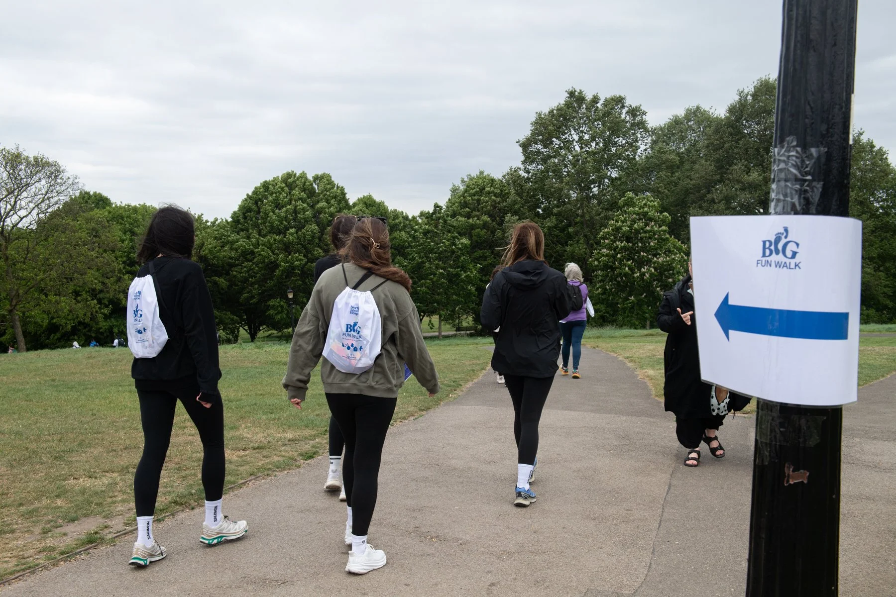 Group of people walking on a park path during a fun walk event, with trees and overcast sky in the background. A sign with an arrow points the direction of the walk.