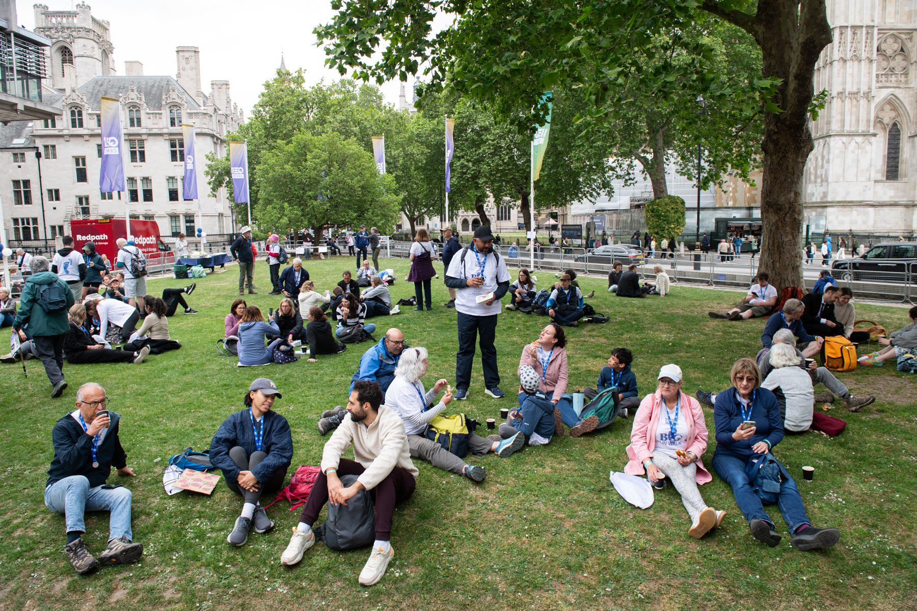 People sitting on the grass in a park, engaging in conversations and using their phones. Some are wearing lanyards, indicating participation in an event. There are trees and historic buildings in the background, with a mix of casual and outdoor cloth