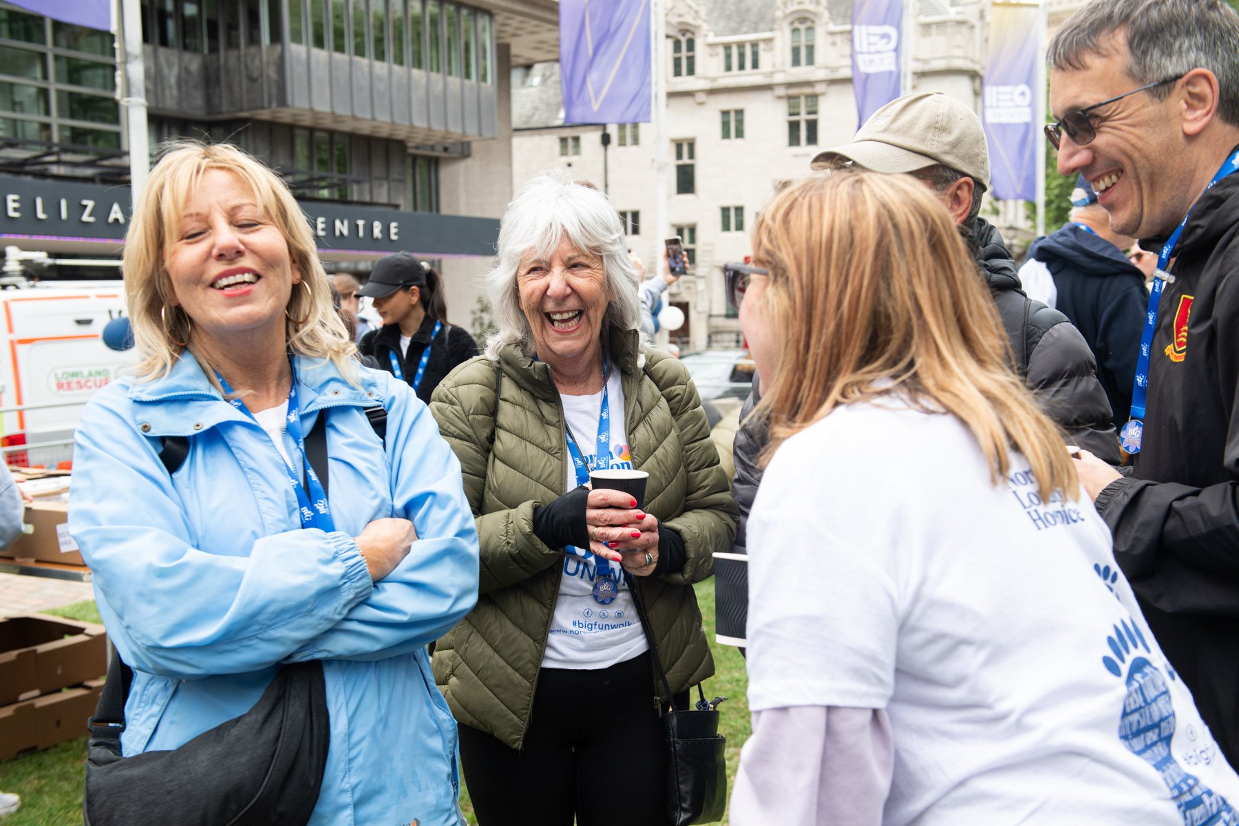 Group of people smiling and talking at an outdoor event, some wearing lanyards and casual jackets, with tents and buildings in the background.