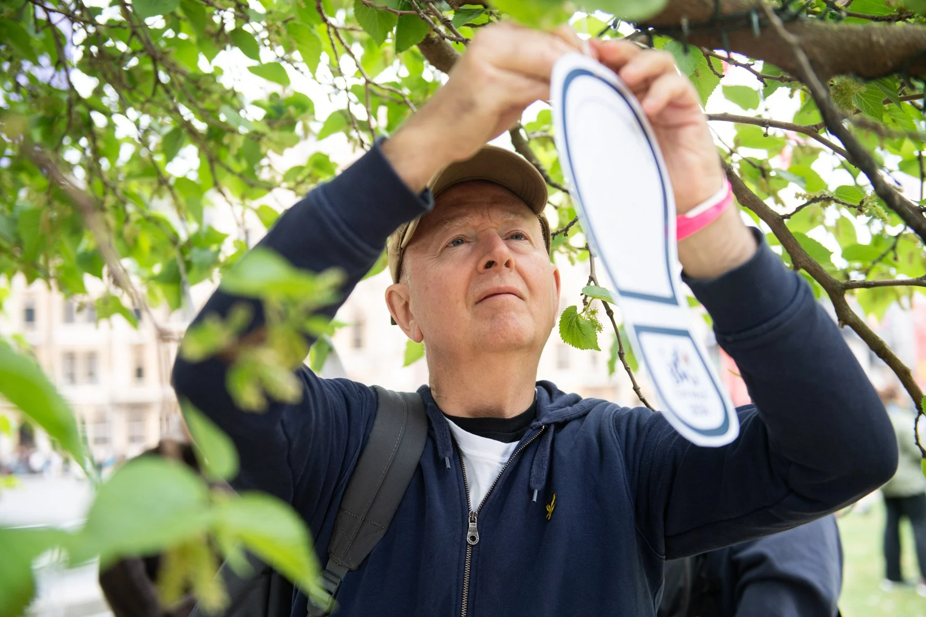 A man with a beige cap and dark jacket carefully examines a plant in a park or urban area, holding a holographic or digital device.