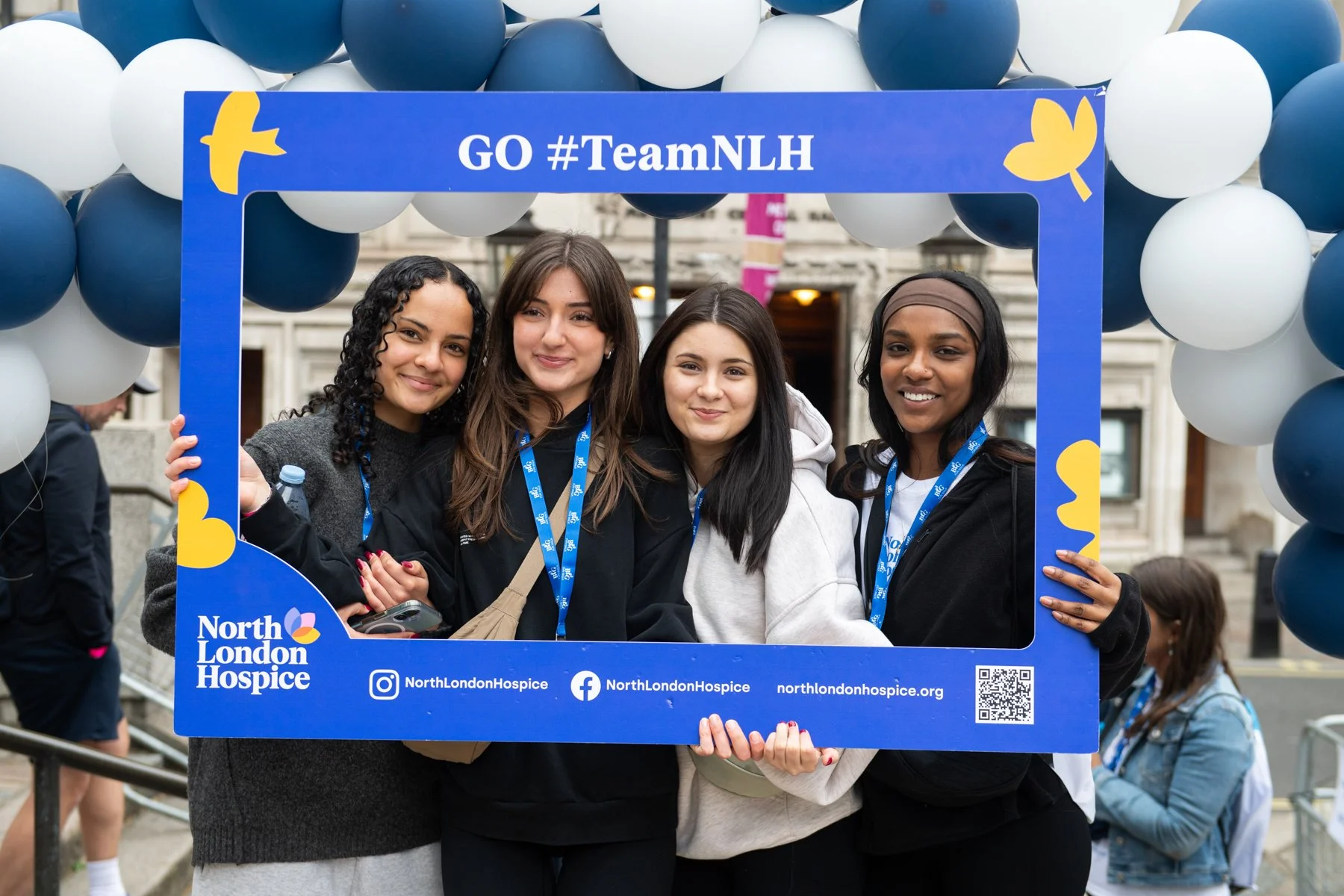 Four young women smiling and posing behind a blue North London Hospice photo frame decorated with balloons, at an outdoor event.