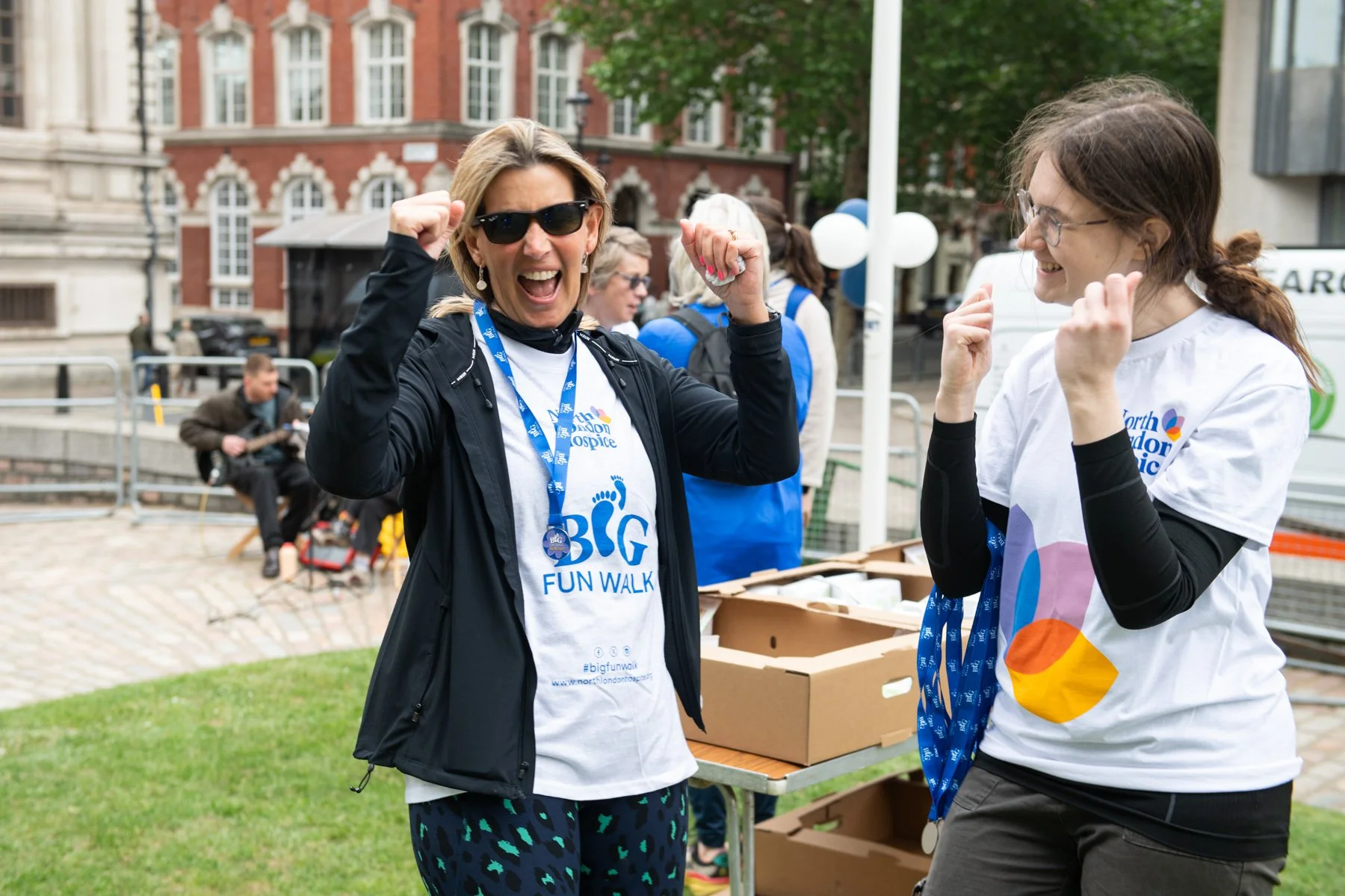 Two women celebrating at a charity fun walk event outdoors, wearing event t-shirts and medals with lanyards, smiling and raising their fists in victory.