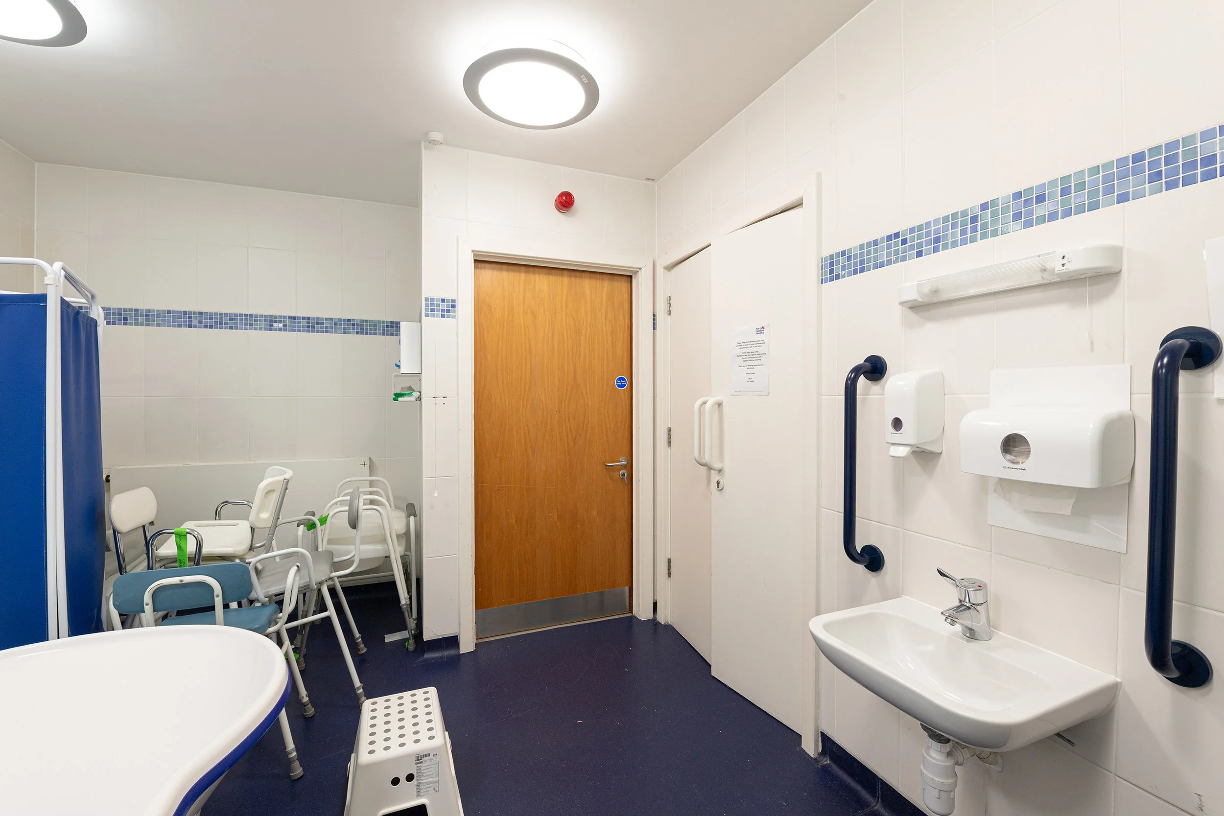 Hospital bathroom with multiple white chairs stacked against the wall, a blue privacy curtain, a sink, and accessibility features including grab bars.