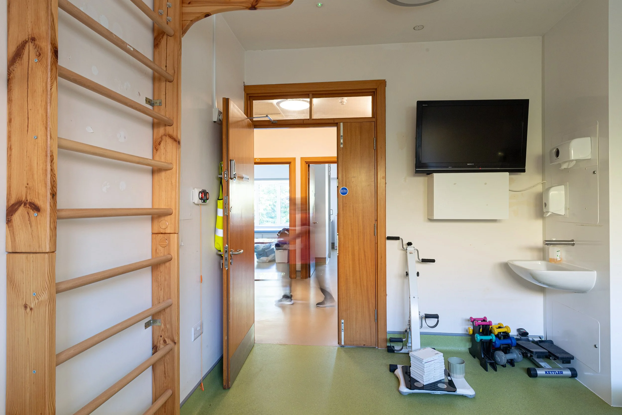 Interior view of a hospital or healthcare facility hallway with beige flooring, white walls, wooden door frames, framed artwork on the walls, ceiling lights, with a person walking away and a reception desk visible in the background.