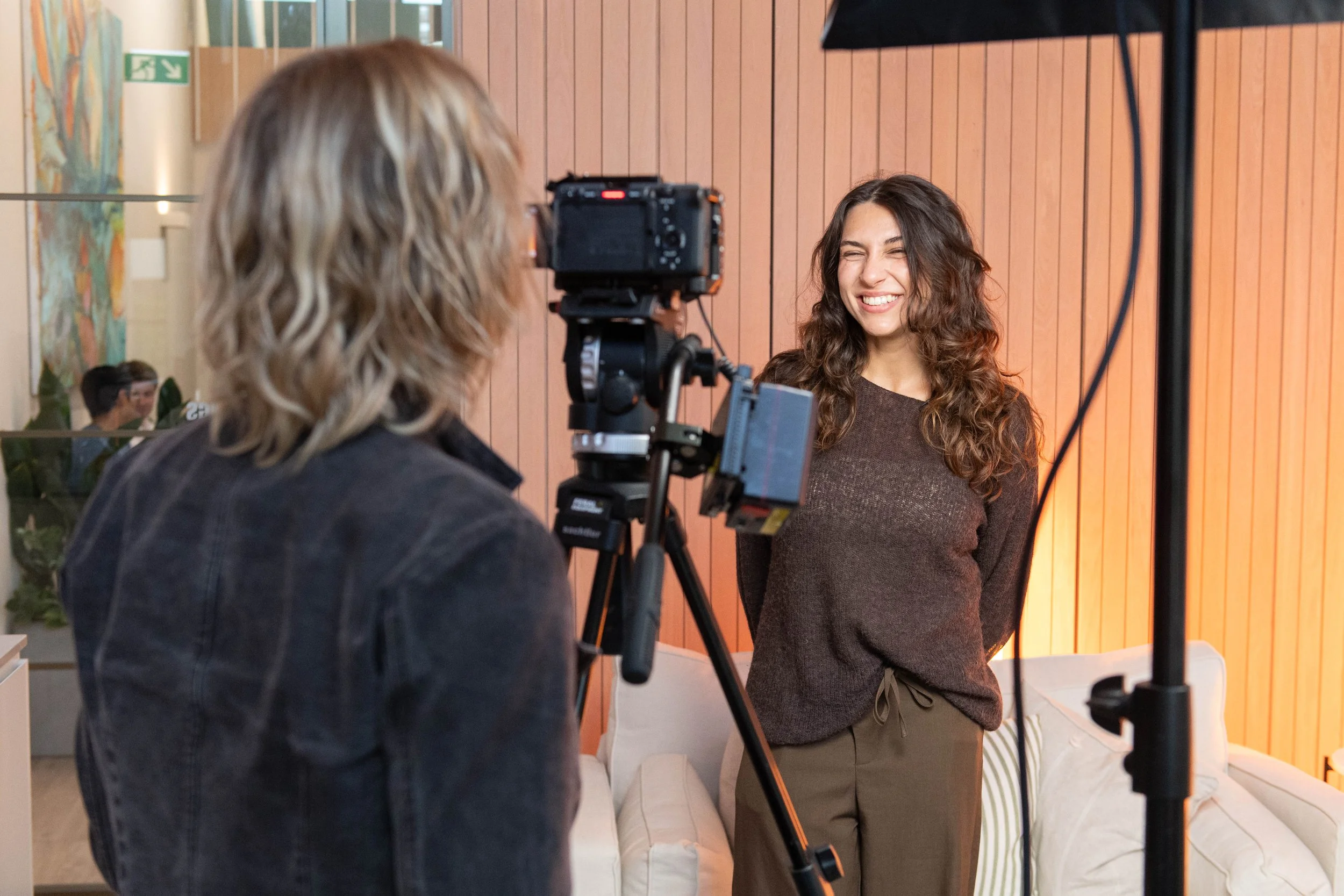 A woman with long curly brown hair smiling and posing for a photo during a photoshoot, in an indoor setting with orange wood-paneled walls.