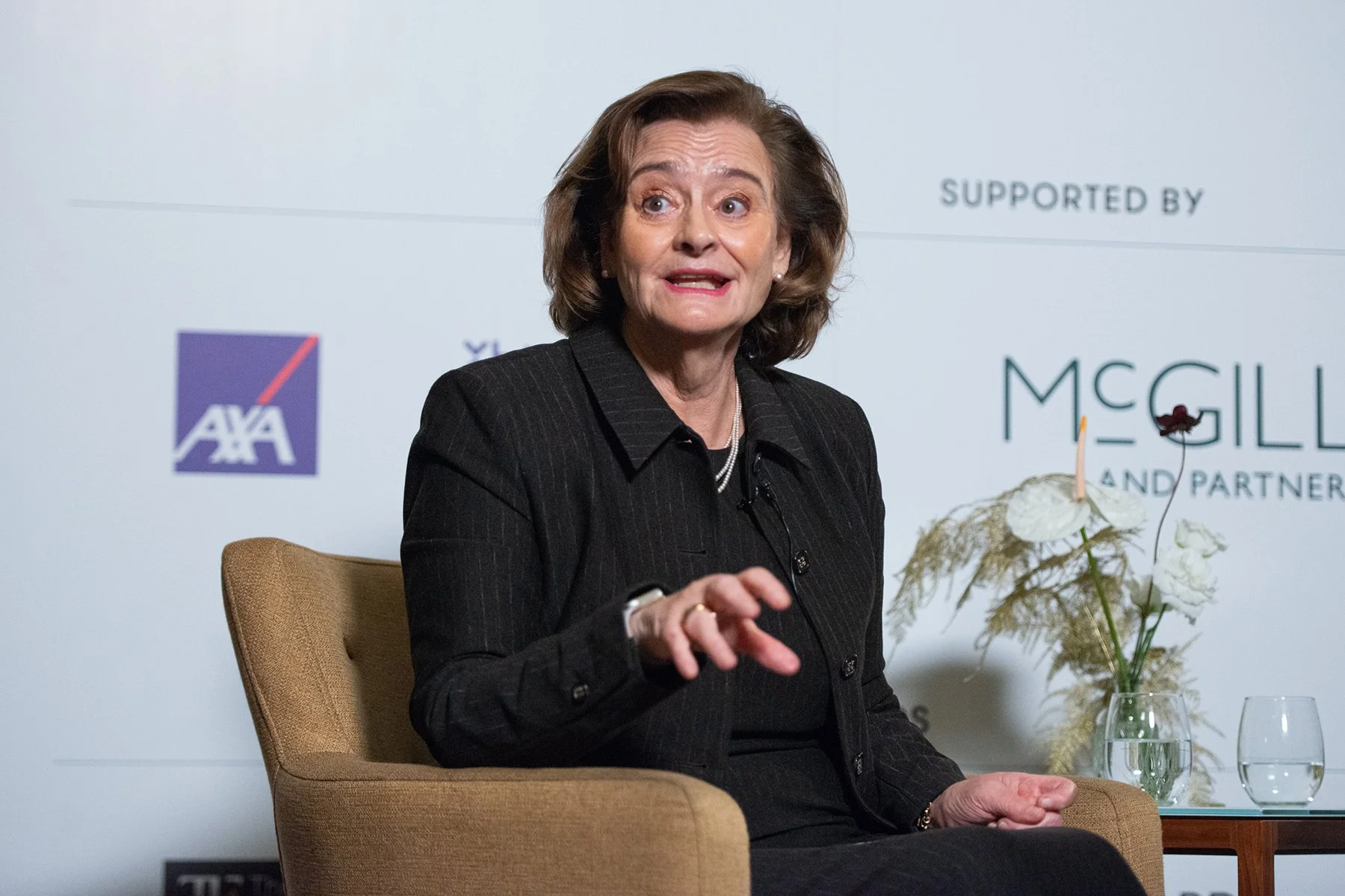 A woman with shoulder-length brunette hair, wearing a black pinstripe blazer, speaking during a panel discussion. She is seated in a beige chair with a bouquet of white flowers and glasses of water on a small table beside her. The background features